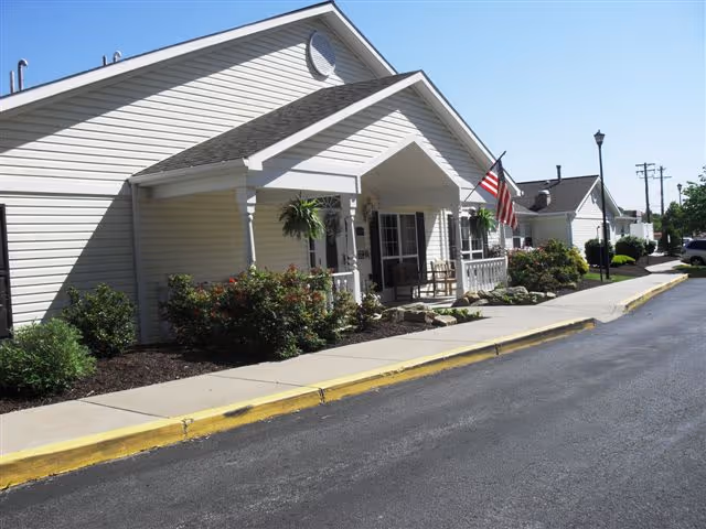 Exterior view of a single-story building with beige siding and a covered porch. The porch has hanging plants, a rocking chair, and an American flag mounted on a pole. There are shrubs and landscaping along the sidewalk, and a paved road runs alongside the building under a clear blue sky.