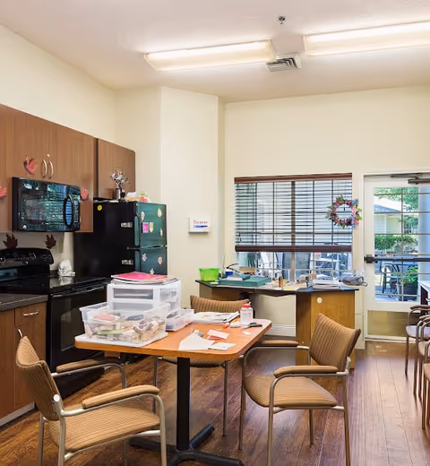 A kitchen area in an assisted living facility with wooden cabinets, a black microwave, stove, and refrigerator. A wooden table with four brown chairs surrounds it, and the table has various plastic containers and papers on it. There is a window with blinds and a door leading outside, decorated with a wreath. The room has wooden flooring and fluorescent ceiling lights.
