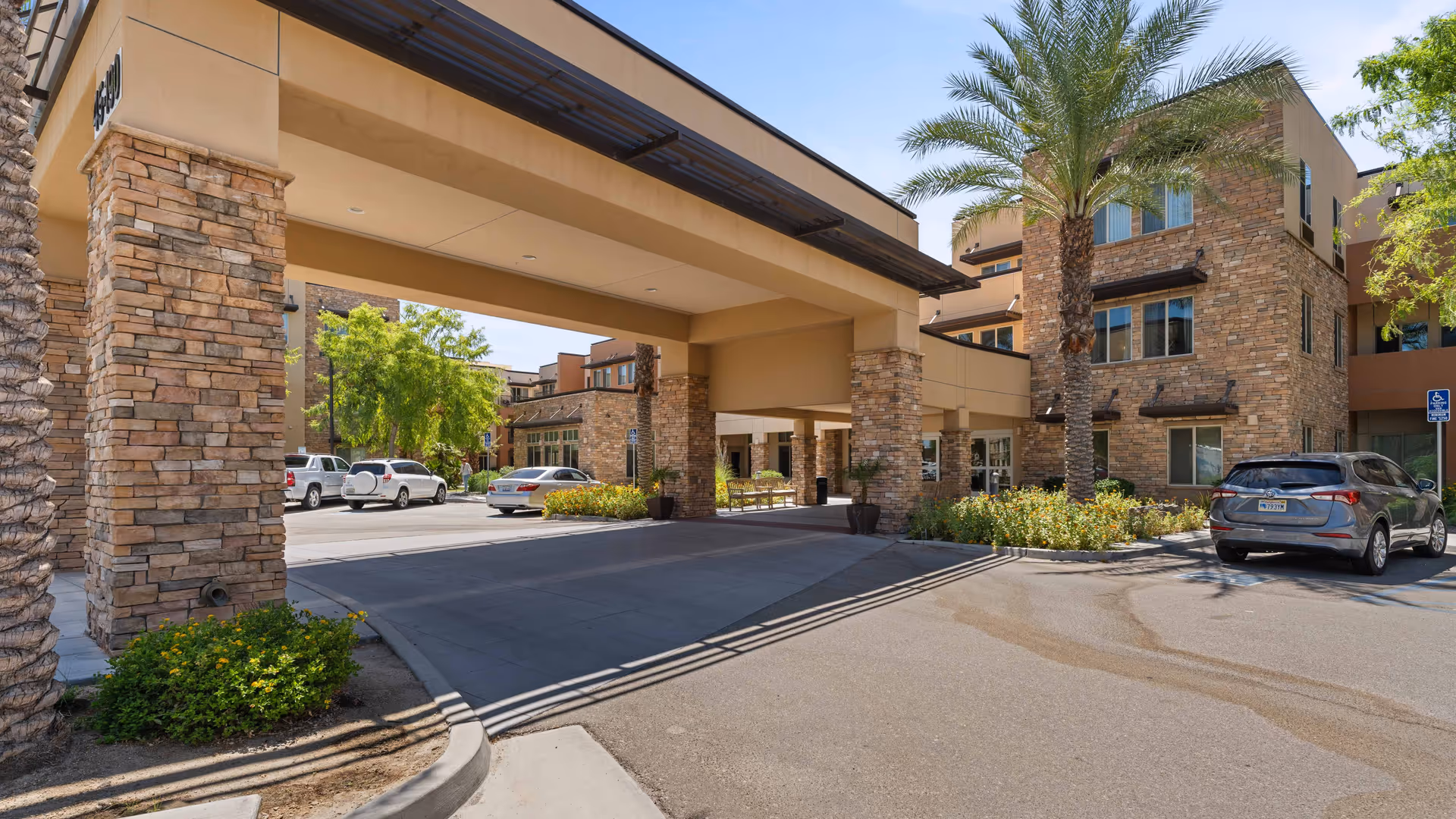 Exterior view of The Palms At La Quinta Gracious Retirement Living facility showing a covered entrance with stone pillars, palm trees, parked cars, and a clear blue sky.