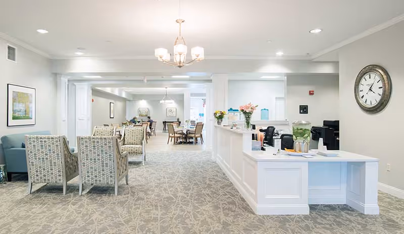 Bright open common area with a reception desk to the right and seating and dining tables beyond in a senior living facility.