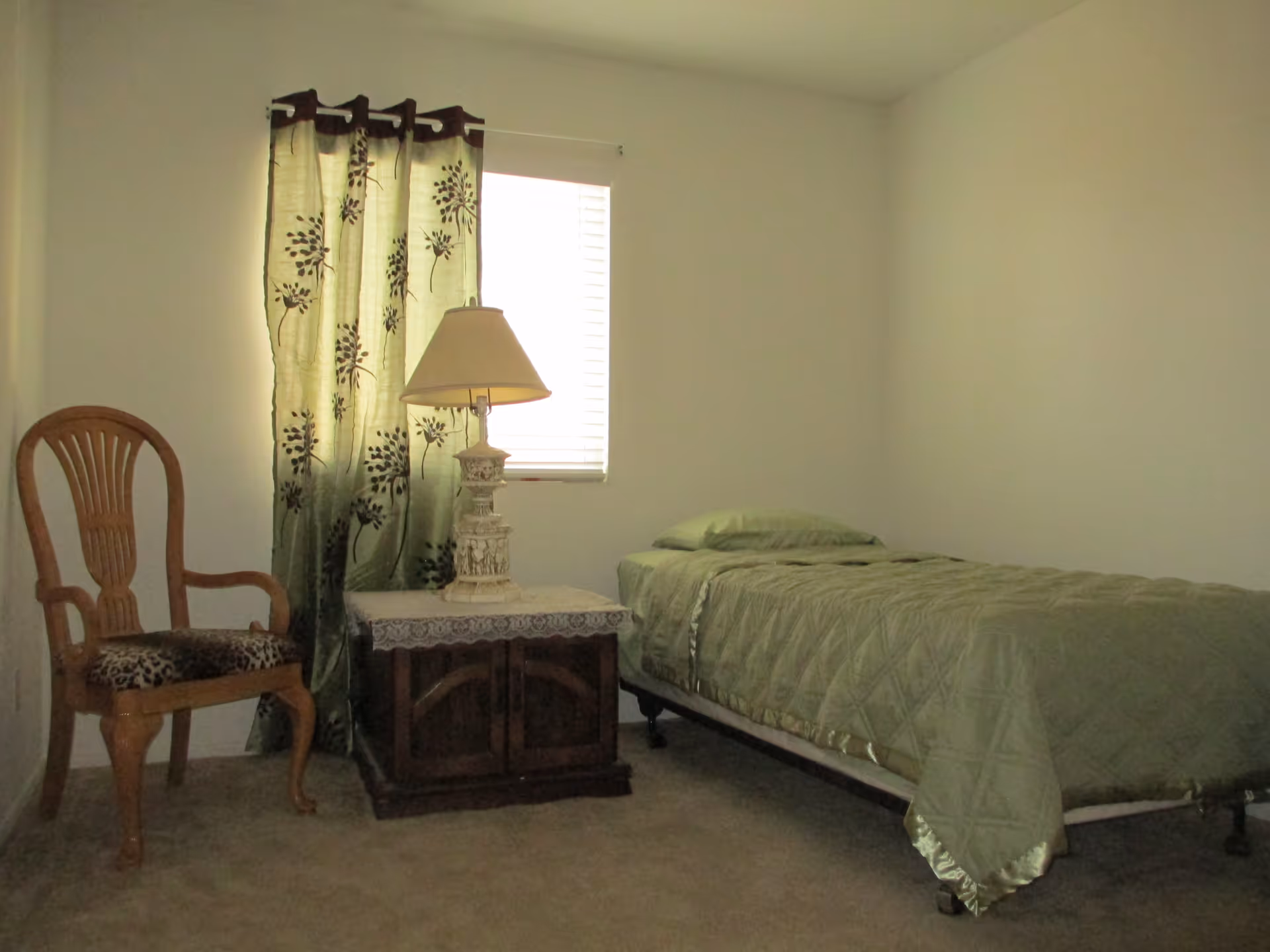 Simple bedroom with a single bed, a wooden chair, a bedside table with a lamp, and green curtains over a window.