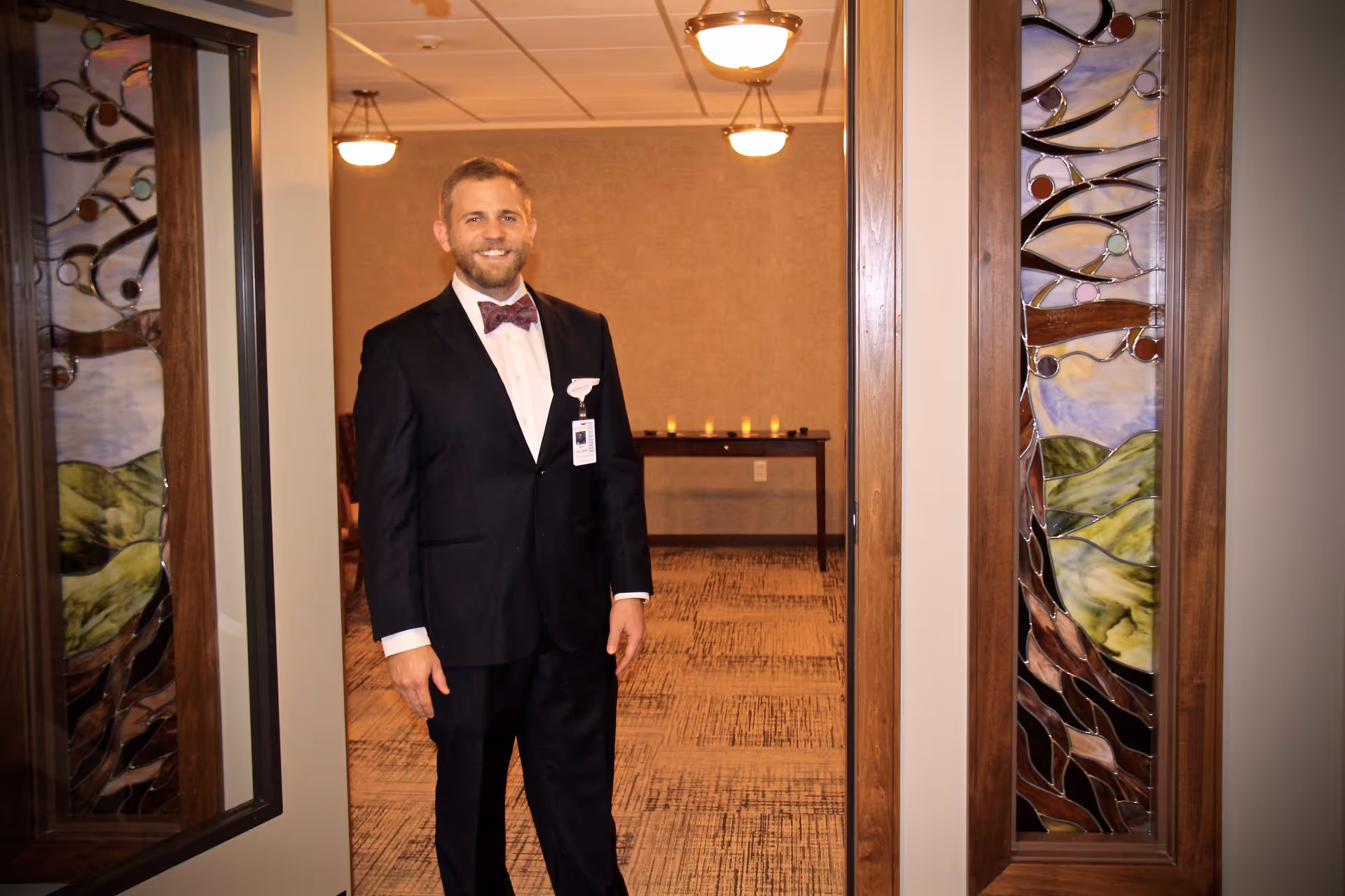 A man in a black suit with a bow tie and name badge stands smiling in a doorway. The doorway features decorative stained glass panels on either side. Behind him is a room with a table holding several lit candles and warm lighting fixtures on the ceiling.