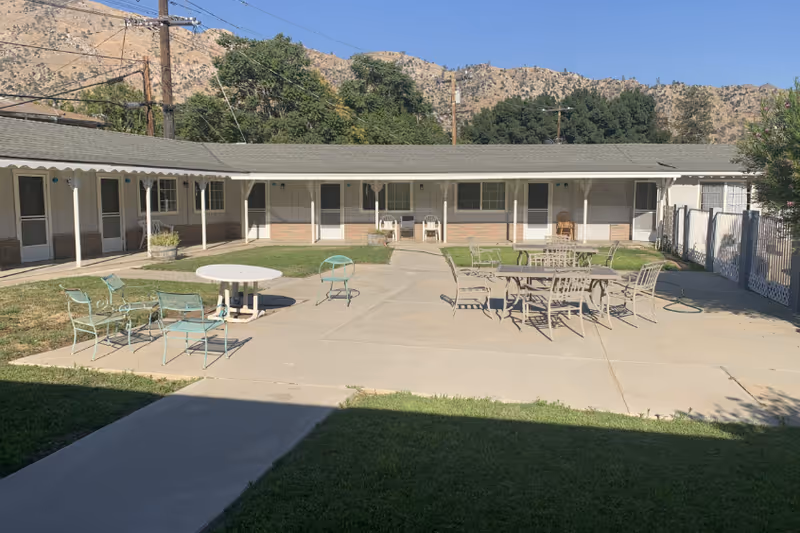 Courtyard of a single-story assisted living facility with patio tables and chairs, rooms opening onto a shaded walkway and hills in the background.