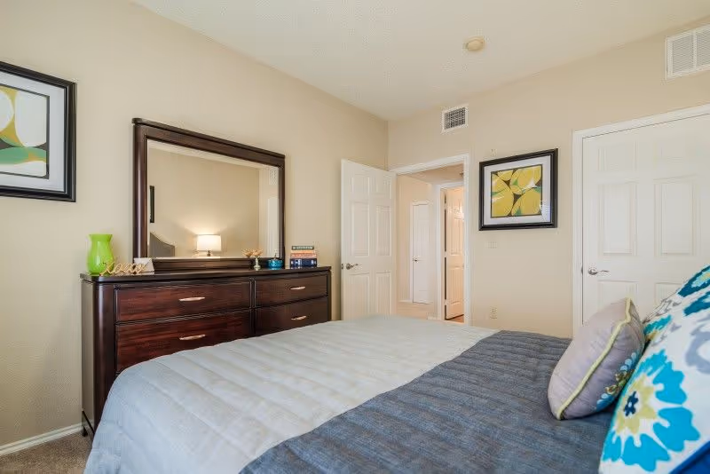 A bedroom with a neatly made bed featuring a blue and gray comforter and decorative pillows. There is a dark wooden dresser with a large mirror above it, decorated with a green vase, books, and small decorative items. The walls are beige with two framed abstract art pieces. Two white doors are visible, one open leading to a hallway and the other closed.
