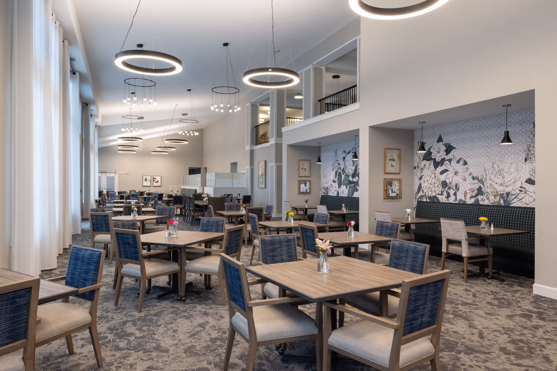 A spacious dining room with multiple square wooden tables and chairs with blue backs and beige seats. Each table has a small vase with flowers. The room features modern circular pendant lights hanging from a high ceiling, large windows with white curtains on the left, and a floral patterned wall with framed pictures on the right. The carpet has a subtle pattern, and the overall decor is modern and inviting.