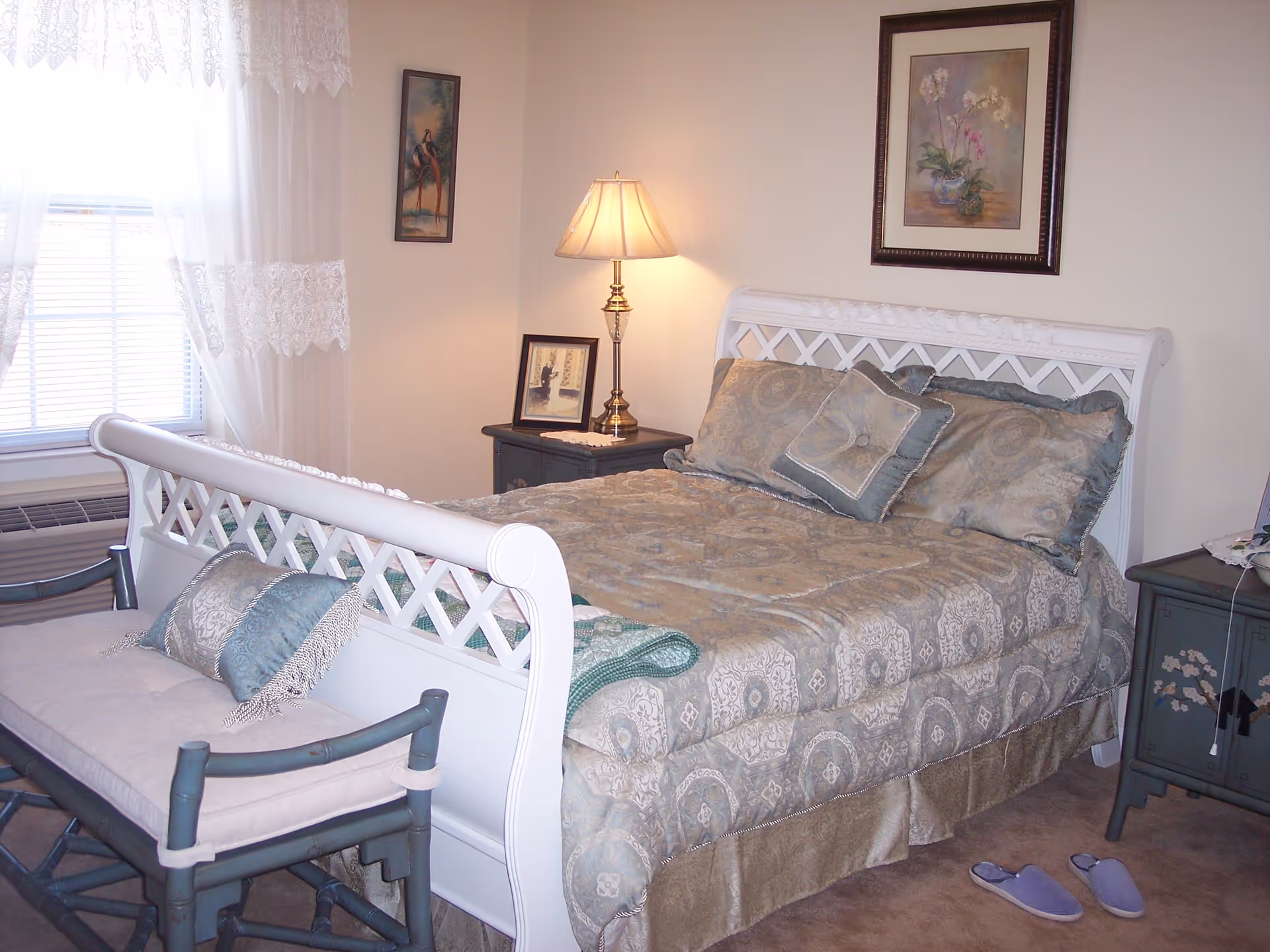 A cozy bedroom with a white wooden bed frame and a patterned bedspread in shades of beige and blue. There are matching pillows on the bed and a small bench with cushions at the foot of the bed. A nightstand with a lamp and framed pictures is beside the bed. The room has beige walls, a window with lace curtains, and a pair of purple slippers on the carpeted floor.