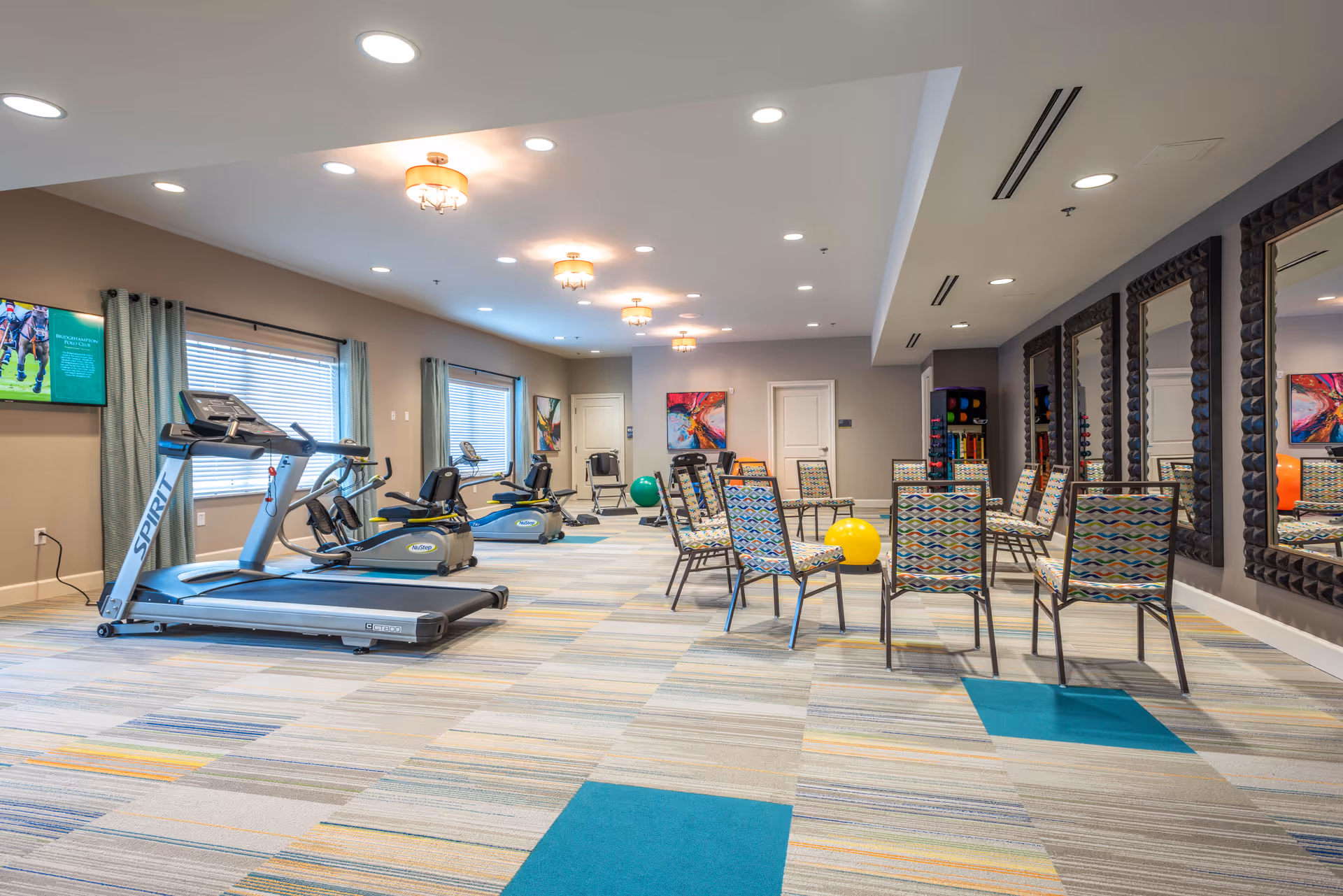 A spacious senior living fitness room with exercise equipment including a treadmill and recumbent bikes on the left side. The right side features a group exercise area with colorful patterned chairs arranged in a circle and large mirrors on the wall. The room has bright overhead lighting, windows with curtains, and colorful abstract artwork on the walls.
