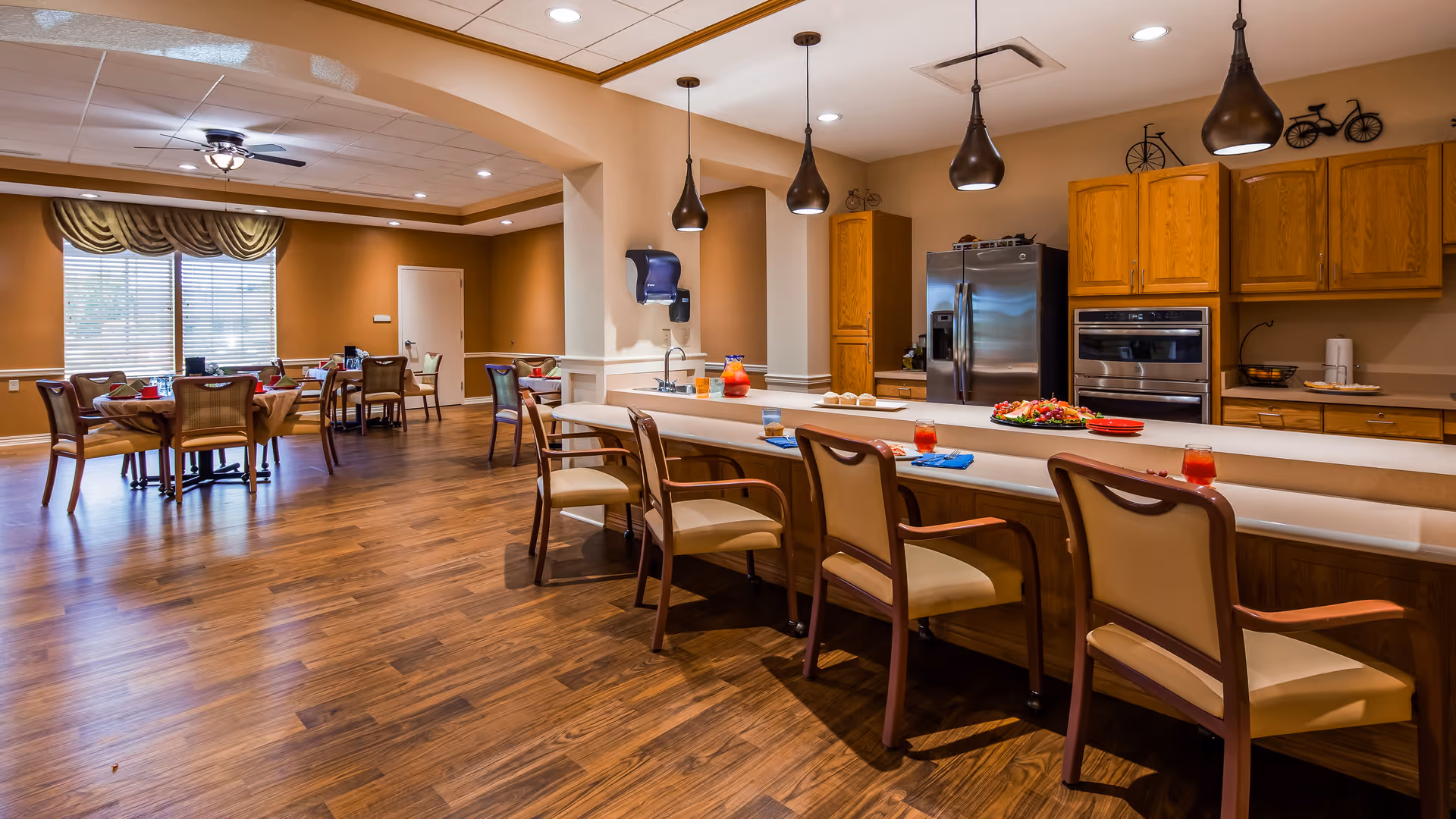 A spacious dining area in a senior living facility featuring a long counter with chairs and place settings, pendant lights hanging above, wooden cabinets, a stainless steel refrigerator, and multiple dining tables with chairs near large windows with blinds and valances.