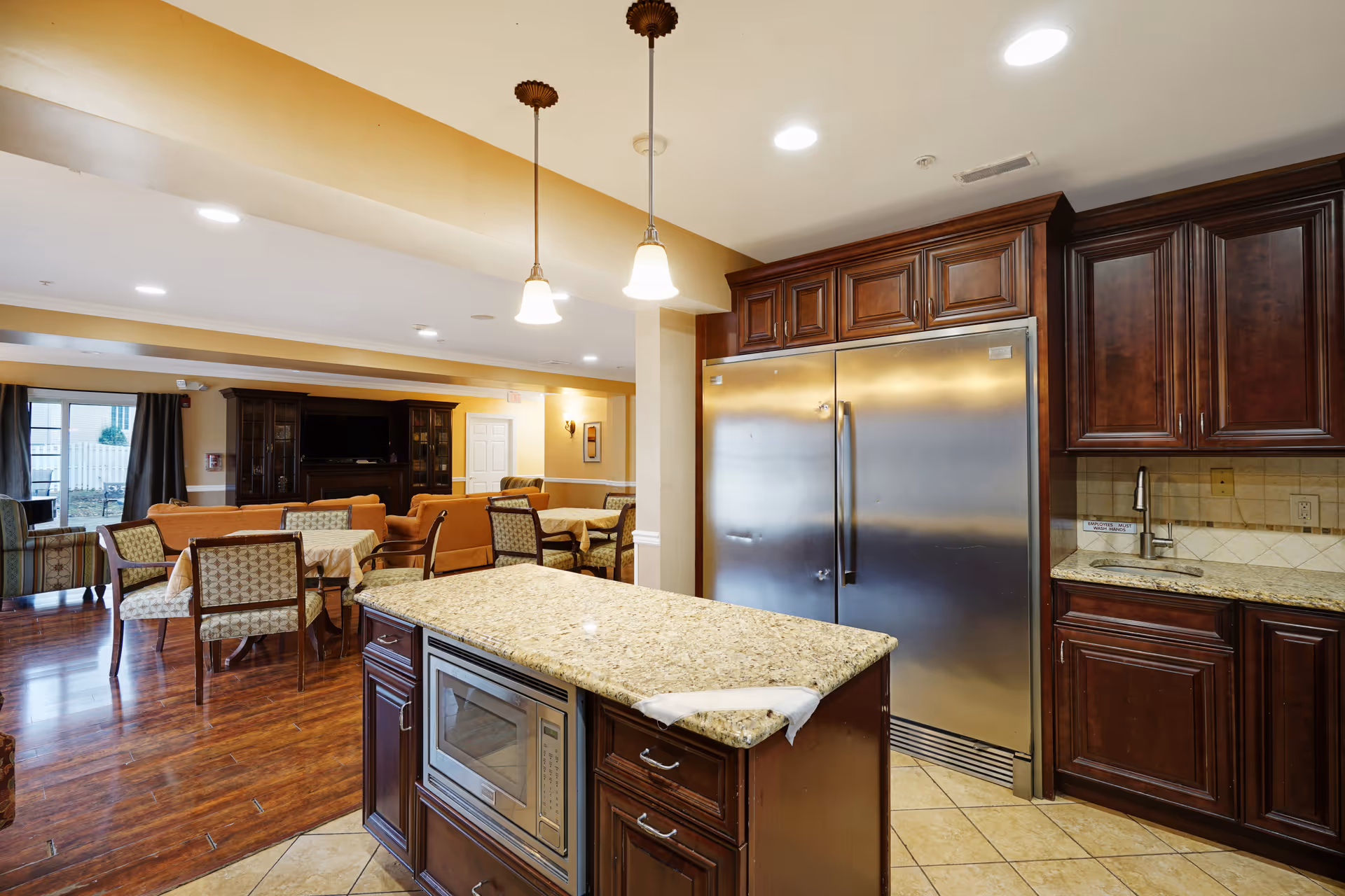 Interior view of a senior living facility kitchen and common area. The kitchen features dark wood cabinets, a large stainless steel refrigerator, a granite countertop island with a built-in microwave, and a small sink. Pendant lights hang above the island. In the background, there is a dining area with tables and chairs, and a living room area with sofas and a large TV cabinet.