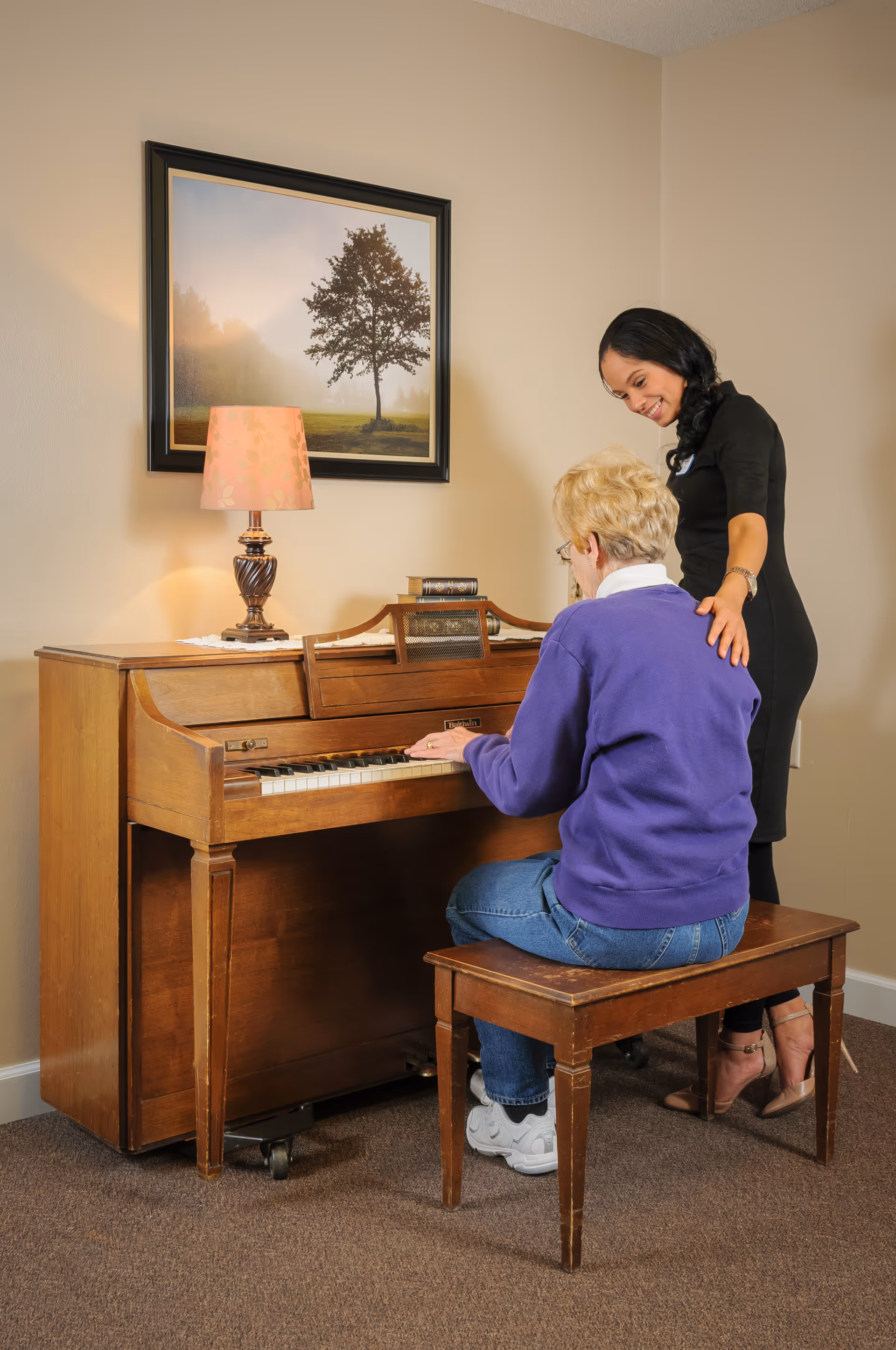 An elderly woman in a purple sweater and jeans sits on a wooden bench playing an upright piano while a younger woman in a black dress stands beside her, smiling and gently touching her shoulder. A table lamp and a framed picture of a tree are on the piano against a beige wall.