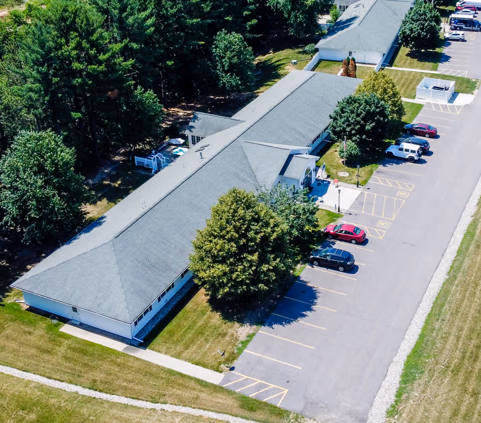 Aerial view of a long, single-story building with a gray roof surrounded by trees and a parking lot with several cars parked. The building is situated in a green area with grass and a sidewalk leading to the entrance.