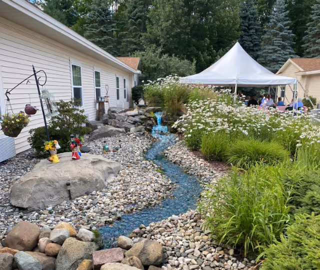 Outdoor garden area at Tawas Village featuring a small artificial stream with blue water flowing over rocks, surrounded by white flowers and green plants. A white canopy tent is set up in the background with people seated underneath. The side of a light-colored building is visible on the left.