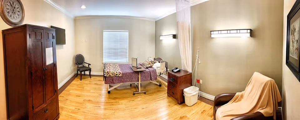 A rehabilitation center room with a hospital bed covered in a purple blanket and floral pillows, a wooden cabinet, a small wooden nightstand with a phone and trash bin, a chair draped with a beige cloth, a wall-mounted TV, a window with blinds, and light fixtures on the walls. The floor is wooden and the walls are painted light beige.