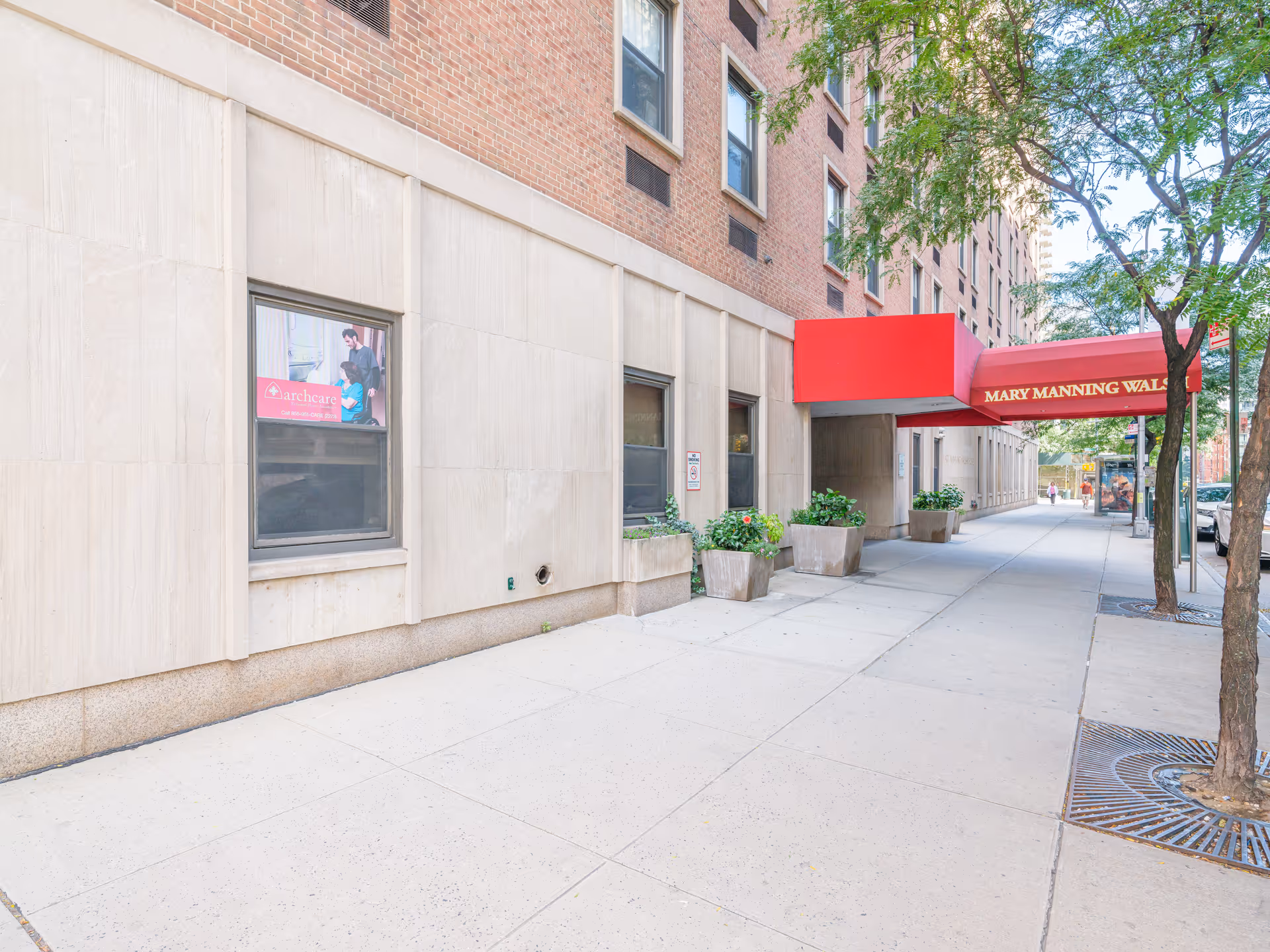 Sidewalk view of the Mary Manning Walsh Nursing Home building exterior with a red canopy over the entrance. There are several large planters with green plants along the building wall and trees lining the sidewalk.