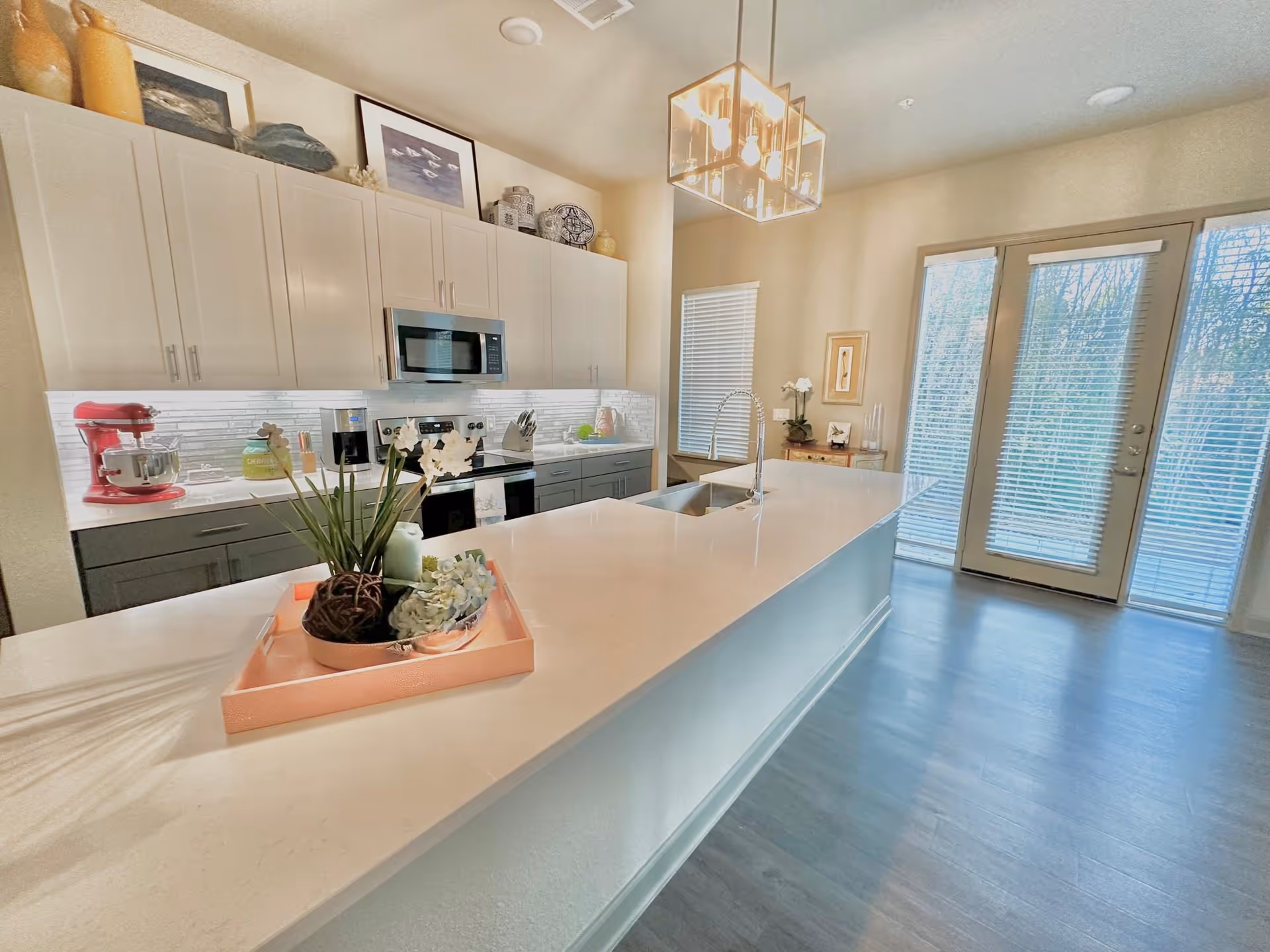 Bright modern kitchen with white countertops and cabinetry, a large island with a sink, a red stand mixer, microwave, stove, and decorative items on the counter and above the cabinets. There are large glass doors with blinds letting in natural light.