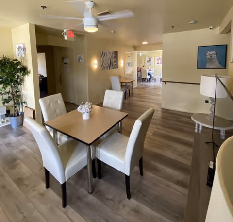A bright and clean common area in an assisted living facility with a square table surrounded by four white cushioned chairs. The room has light-colored walls and wood flooring. There is a ceiling fan with a light, framed animal artwork on the walls, a potted plant, and a lamp on a side table. In the background, another dining area with tables and chairs is visible.