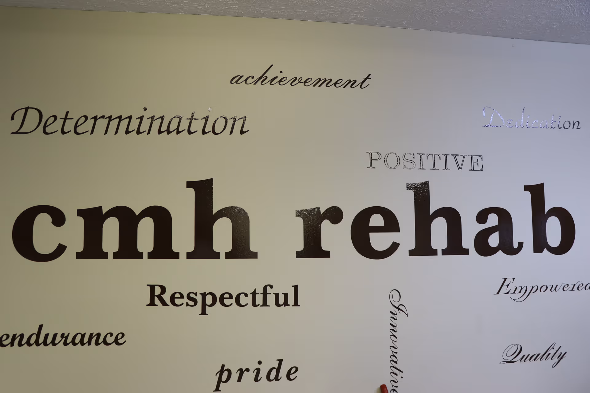 A wall with various motivational words in different fonts and sizes, including 'cmh rehab' prominently in the center, surrounded by words like Determination, achievement, Dedication, POSITIVE, Respectful, Empowered, endurance, pride, Innovation, and Quality.
