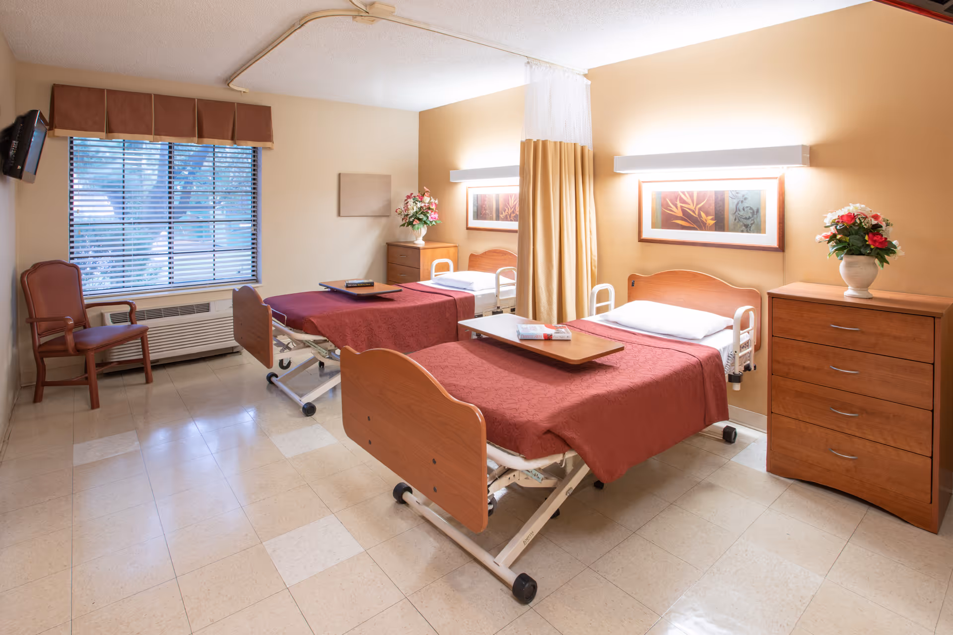 A bright and clean room with two hospital-style beds covered with red bedspreads, each with a wooden overbed table. The room has beige walls, a window with blinds and a brown valance, a wooden chair, two wooden dressers with flower vases on top, framed artwork above the beds, and a ceiling-mounted privacy curtain between the beds.
