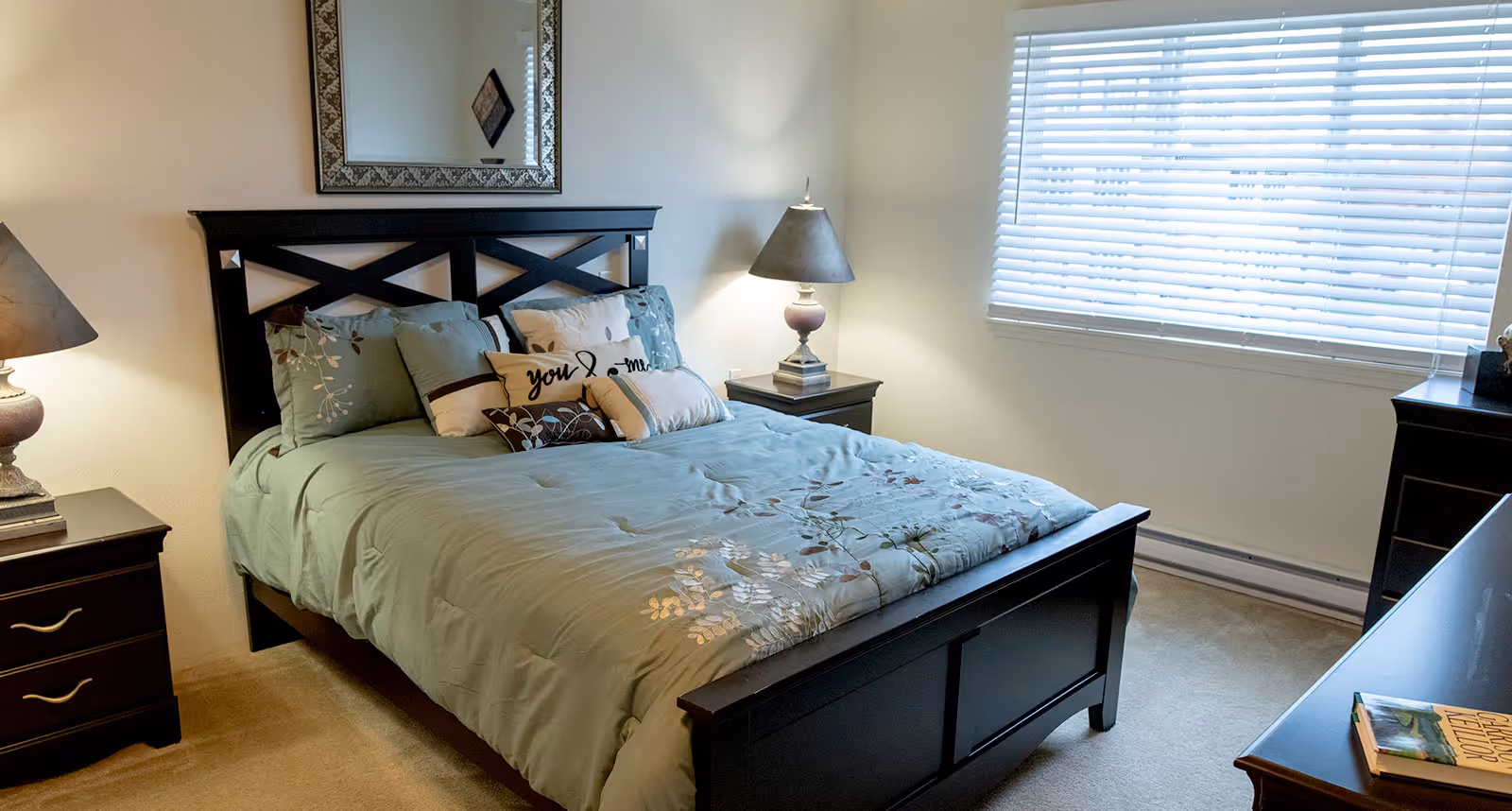 Cozy bedroom with a dark wood bed dressed in a green floral comforter, matching nightstands with lamps, a window with blinds, and a dresser.
