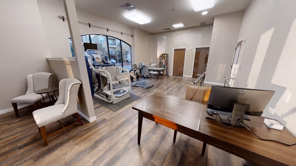 A spacious room with wooden flooring featuring exercise equipment including a treadmill and stationary bike, a wooden desk with a computer and a clear protective screen, and two upholstered chairs with a small round table between them. Large windows allow natural light to fill the room.