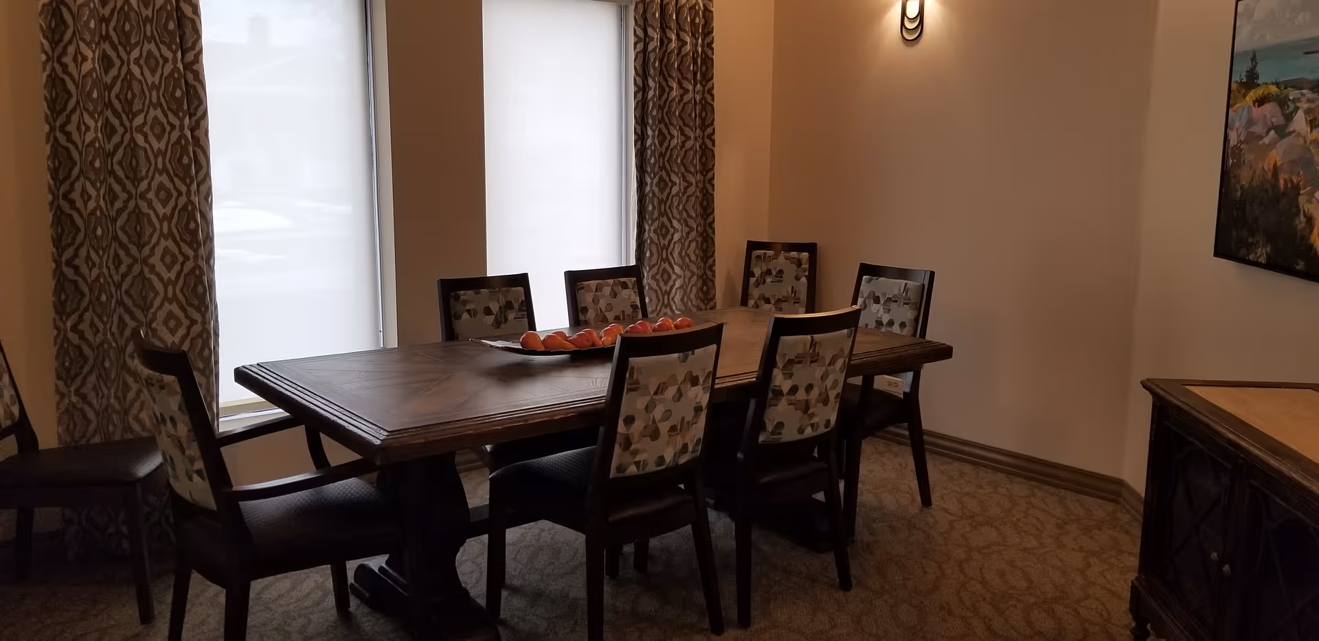A dining room with a rectangular wooden table surrounded by six chairs with patterned upholstery. The room has two large windows with white blinds and patterned curtains. There is a decorative bowl with red apples on the table, a wall-mounted light fixture, a painting on the wall, and a wooden sideboard.