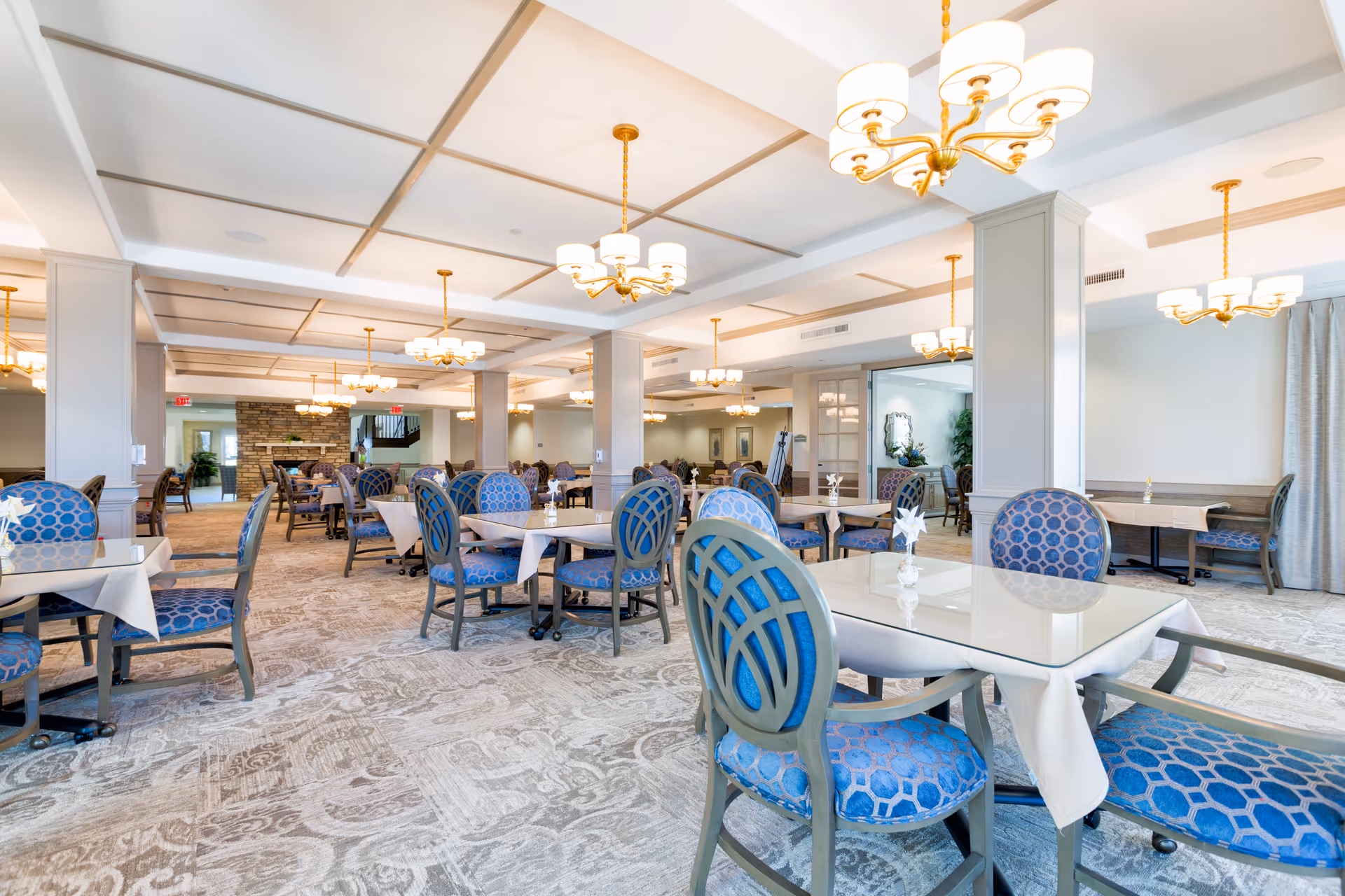 A spacious and well-lit dining room with multiple tables covered with white tablecloths and surrounded by blue patterned chairs. The room features elegant chandeliers hanging from a coffered ceiling, carpeted floors with a subtle pattern, and a stone fireplace in the background. The overall ambiance is bright and inviting.