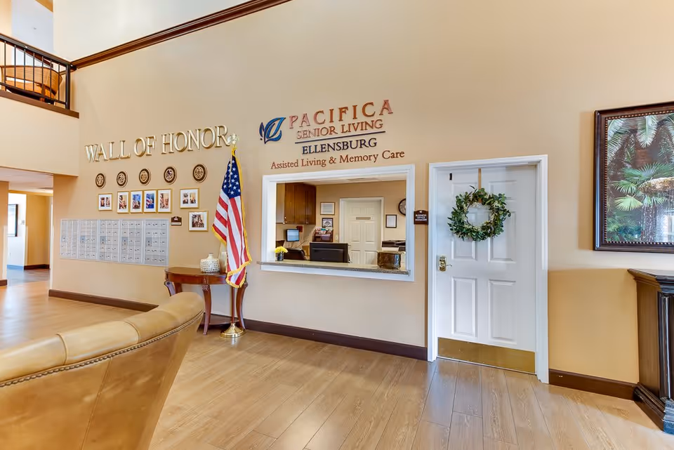 Interior view of the lobby area at Pacifica Senior Living Ellensburg featuring a reception window, a white door with a green wreath, an American flag on a stand, a Wall of Honor with photos and plaques, and a framed tropical-themed painting on the wall.