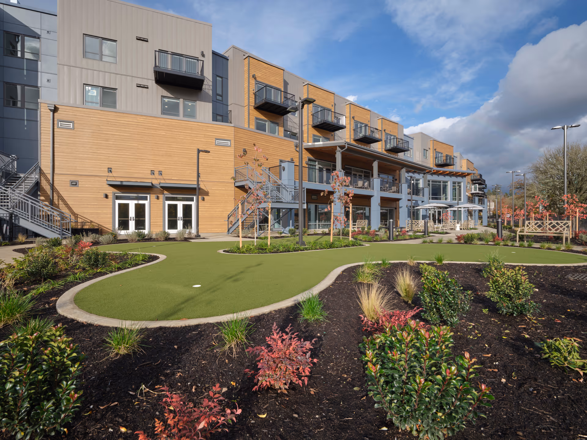 Exterior view of a modern multi-story senior living facility with balconies, large windows, and a landscaped garden area featuring a putting green and various shrubs and small trees under a partly cloudy sky.
