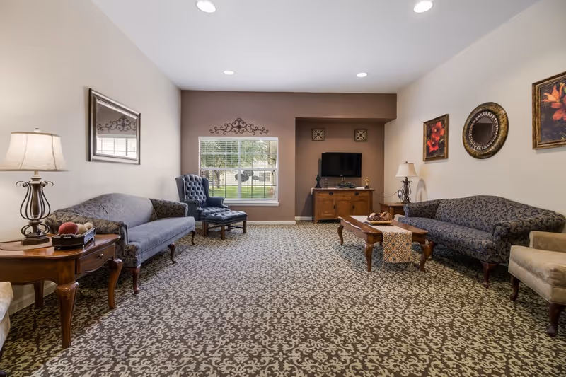 A spacious living room with patterned carpet, two upholstered sofas, a blue armchair, wooden coffee table with decorative items, side tables with lamps, a wall-mounted TV on a wooden cabinet, and framed artwork and a mirror on the walls. A window shows a view of the outdoor greenery.