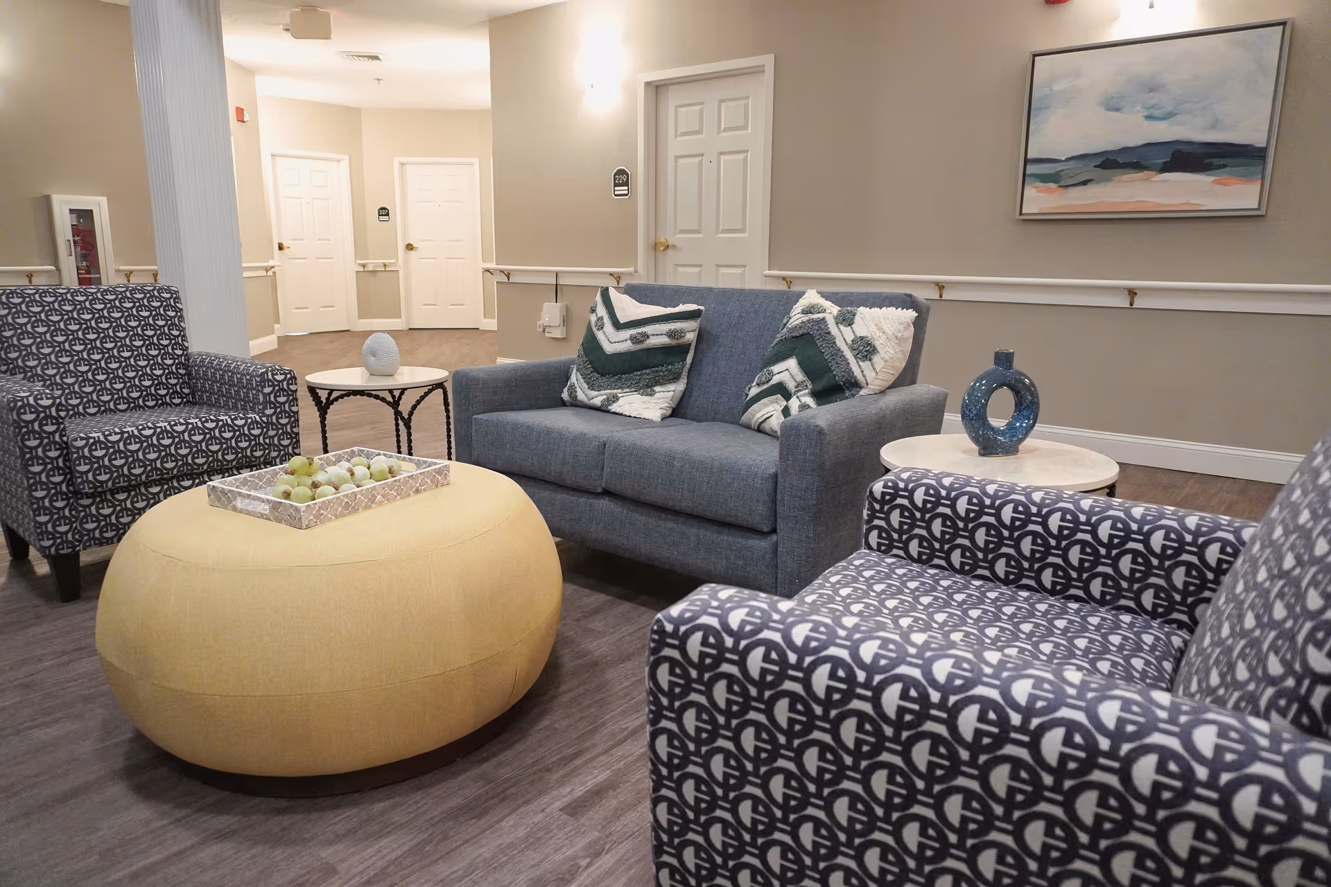 A cozy seating area in an assisted living facility with two patterned armchairs, a blue loveseat with two decorative pillows, a round yellow ottoman with a tray of green grapes, and two small round side tables with decorative objects. The walls are beige with white trim, and there are several white doors and a framed abstract painting on the wall.
