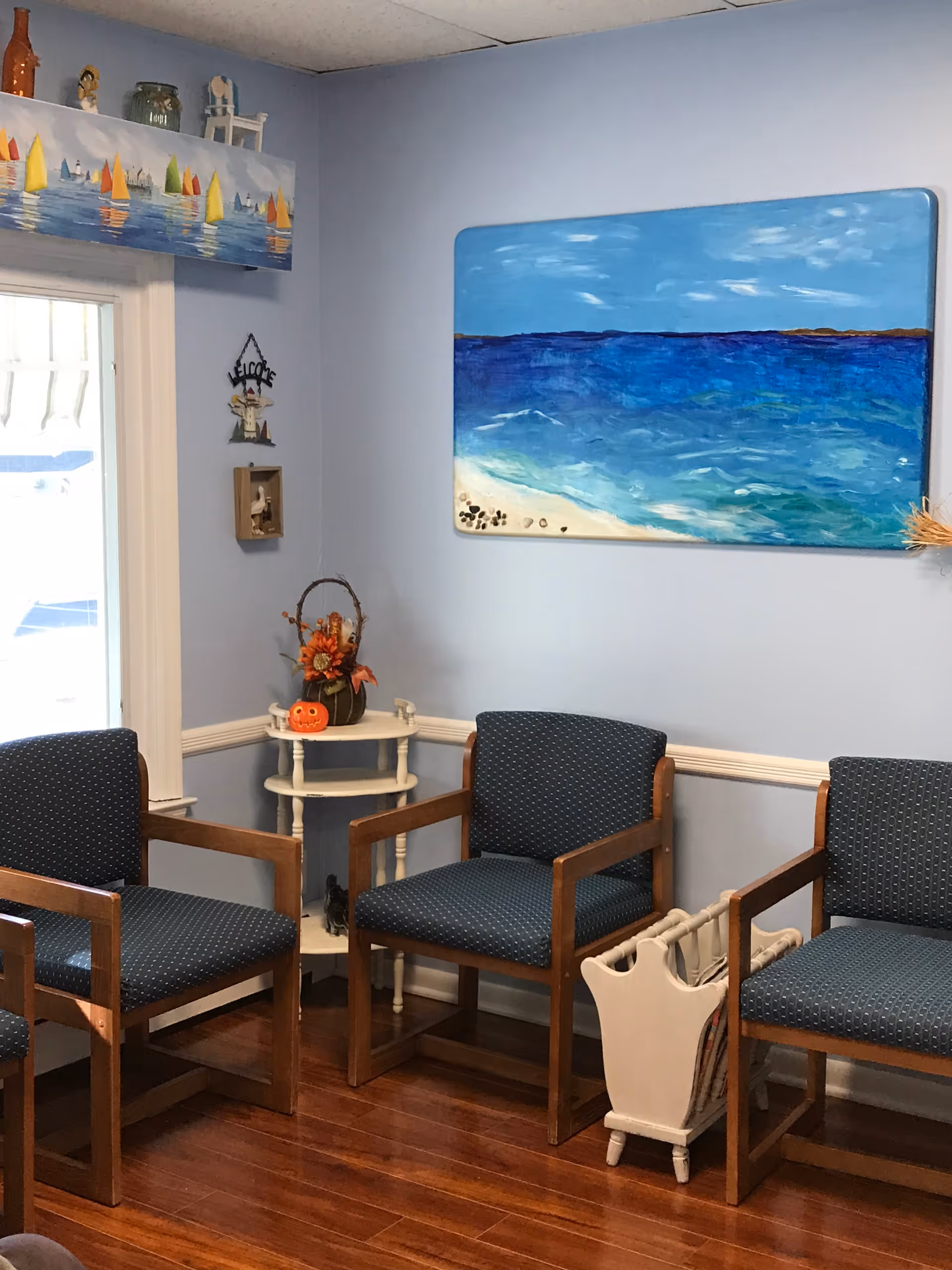 A small waiting area with four wooden chairs upholstered in dark blue fabric with small white dots. The walls are painted light blue and decorated with a large painting of a beach and ocean, a small 'Welcome' sign, and a shelf with small decorative items including a pumpkin and flowers. The floor is wooden, and there is a white magazine rack holding newspapers or magazines between two chairs.