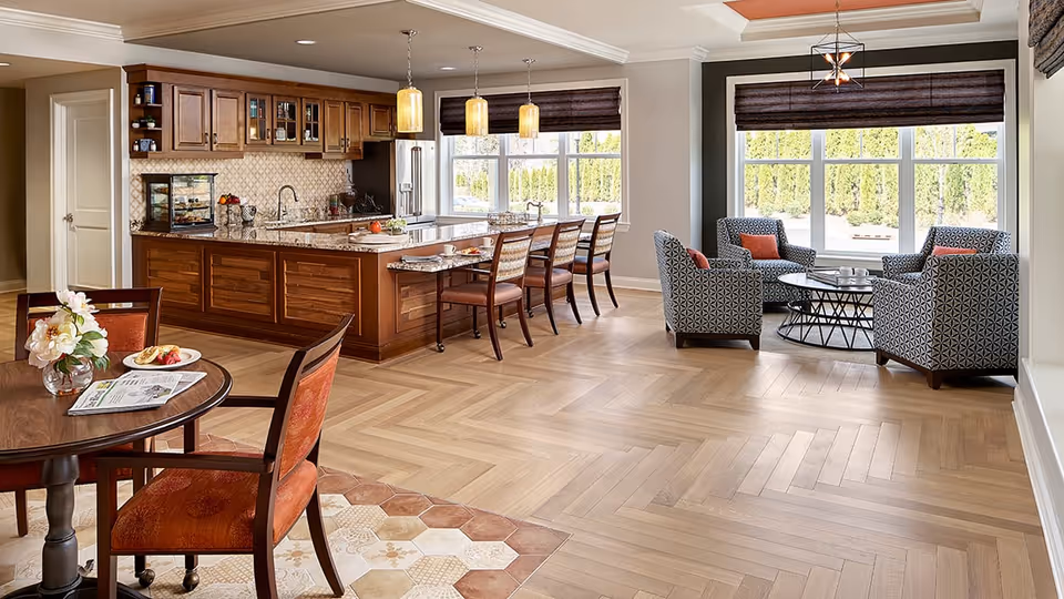 A spacious and well-lit interior area featuring a kitchen with wooden cabinets and a granite countertop island with bar stools. Adjacent to the kitchen is a small dining table with two chairs, and a cozy sitting area with four patterned armchairs arranged around a round coffee table near large windows with dark blinds.