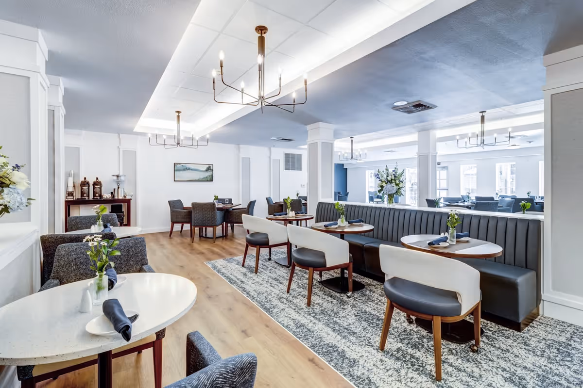 A bright and modern dining room with multiple round tables set with napkins and small flower vases. The room features a long black cushioned bench along one side, paired with white and black chairs. The floor is a combination of wood and a patterned rug. There are several chandeliers hanging from the ceiling, and a sideboard with coffee dispensers and cups is visible in the background.