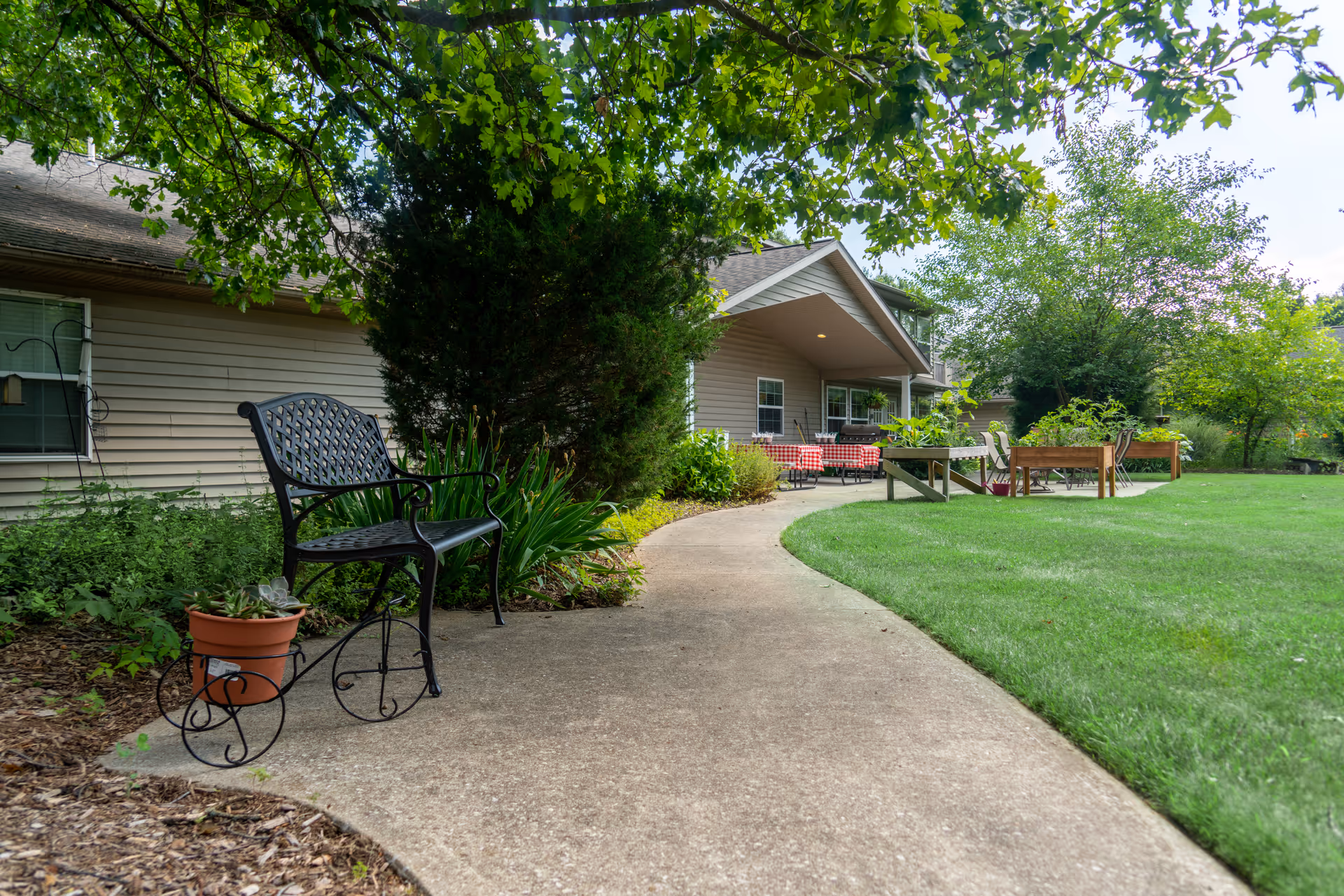 A curved concrete walkway through a grassy yard leads to a patio with chairs beside a single-story building, with a bench and potted plant in the foreground.
