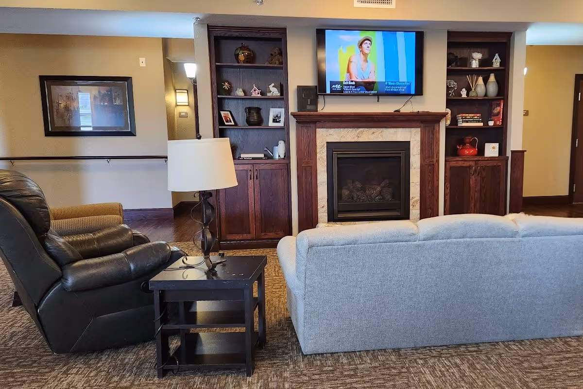 A cozy living room area in a senior living facility featuring a gray sofa and a black recliner chair separated by a small black side table with a lamp. In front of the seating is a fireplace with a wooden mantel and built-in shelves on either side holding decorative items and books. A flat-screen TV is mounted above the fireplace displaying a program. The room has warm lighting and neutral-colored walls and carpet.