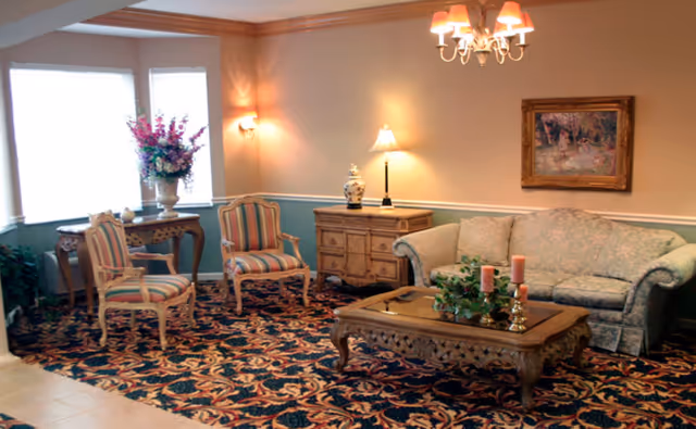 A cozy living room area with a patterned carpet, a floral upholstered sofa, two striped armchairs, a wooden coffee table with candles and greenery, a side table with a lamp and decorative vase, a console table with a large flower arrangement, and a framed painting on the wall. The room is softly lit by wall sconces and a chandelier.