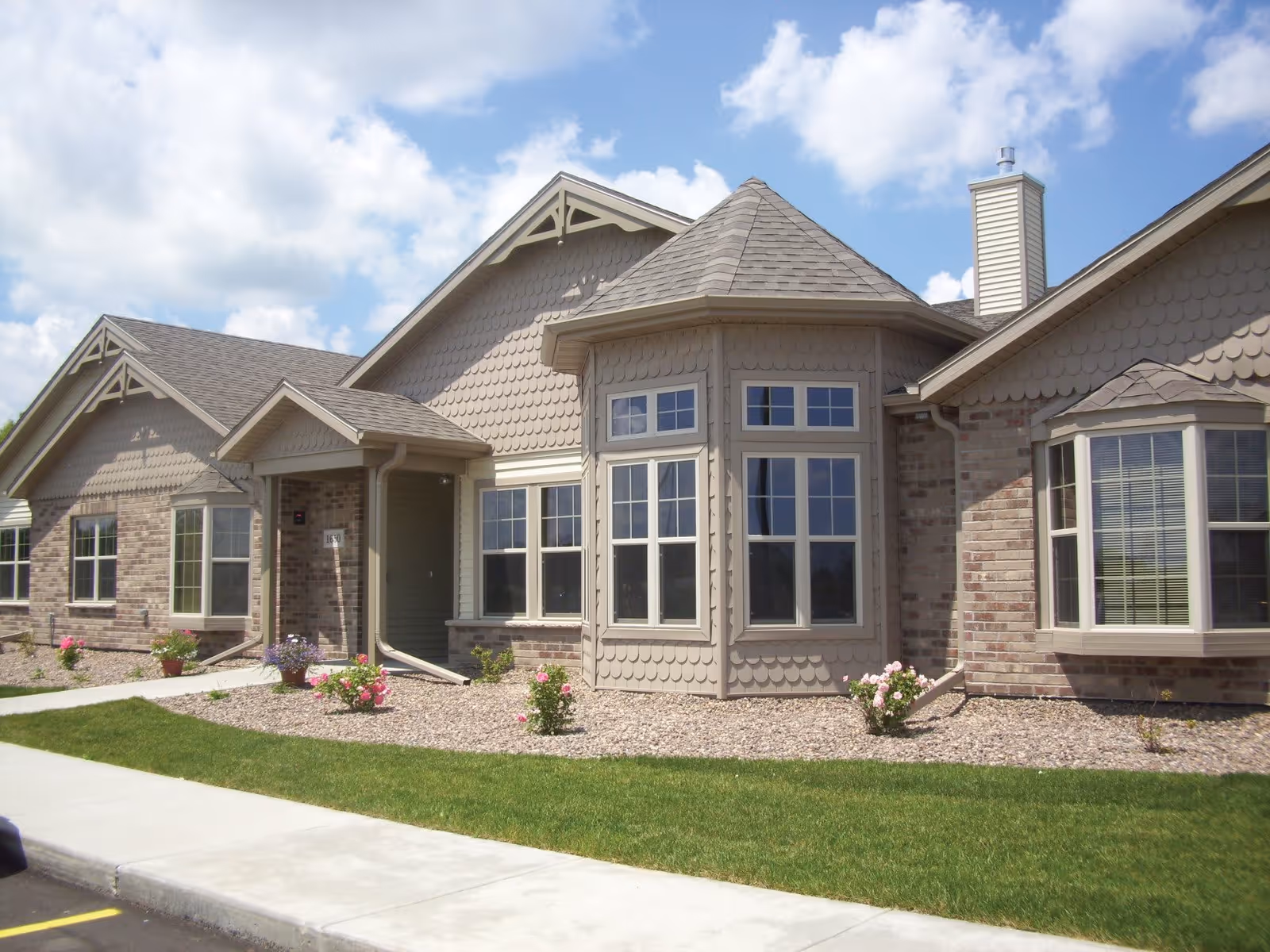 Exterior view of a single-story assisted living facility building with beige siding and brick accents, multiple windows including a bay window, a small covered entrance, landscaped with green grass and flower beds, under a partly cloudy sky.