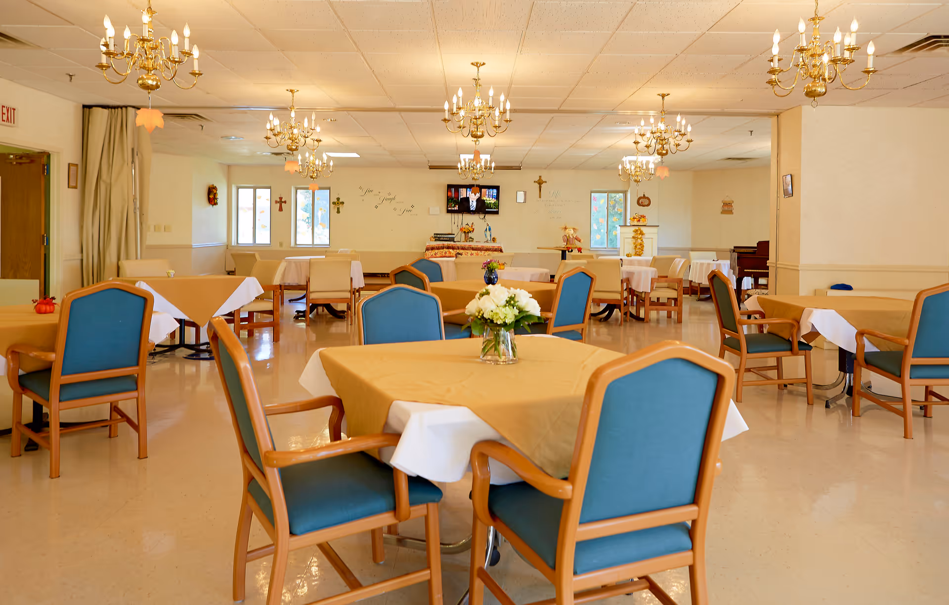 A spacious dining room with multiple tables covered in beige tablecloths and white underlays, surrounded by wooden chairs with teal cushions. The room is well-lit with several ornate chandeliers hanging from the ceiling. There are windows on the far wall, religious crosses, and a television mounted on the wall. The floor is a light beige color, and there are floral centerpieces on the tables.