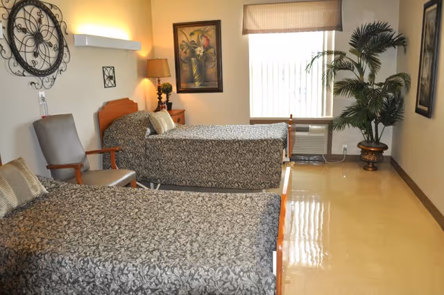 A room with two single beds covered in patterned bedspreads, a gray armchair, a nightstand with a lamp, a large decorative wall clock, framed artwork, a window with blinds, an air conditioning unit below the window, and a large potted plant in the corner.