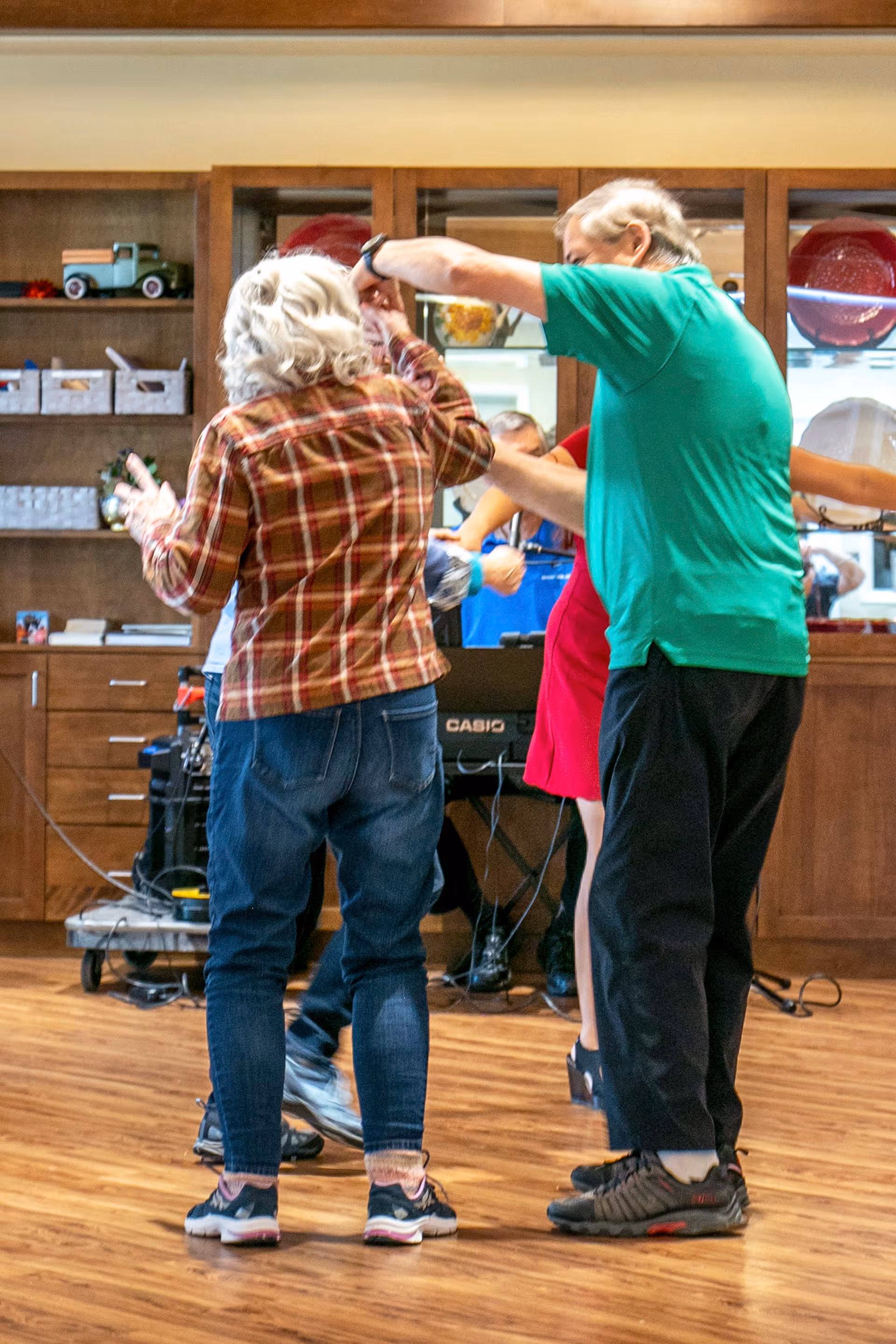 A group of elderly people dancing together in a room with wooden floors and a wooden cabinet in the background. One person is wearing a green shirt and black pants, and another is wearing a plaid shirt and jeans. There is a keyboard and some audio equipment visible behind them.