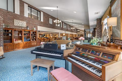 Spacious senior living facility common area with a blue patterned carpet, a wooden organ and a black grand piano in the foreground, comfortable seating areas with sofas and chairs, wooden bookshelves along a brick wall, and chandeliers hanging from the ceiling.