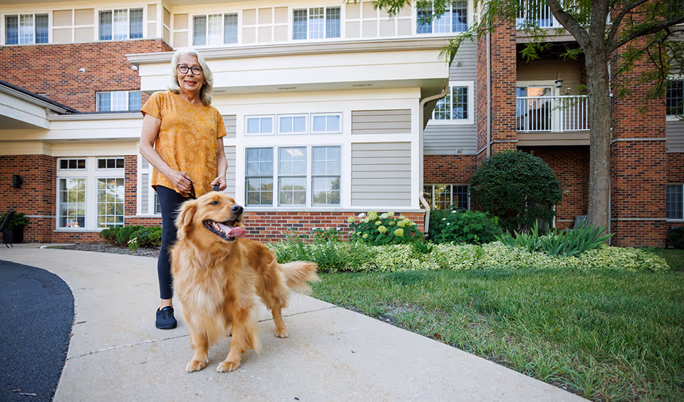 An older woman standing on a sidewalk with a golden retriever in front of a brick senior living building entrance.