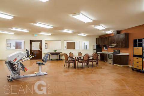 A spacious room with wooden flooring featuring a small kitchen area with dark cabinets, a stove, and a microwave on the right side. In the center, there is a round table with six chairs around it. On the left side, there is a piece of exercise equipment. The room has several ceiling lights and windows on the far wall.
