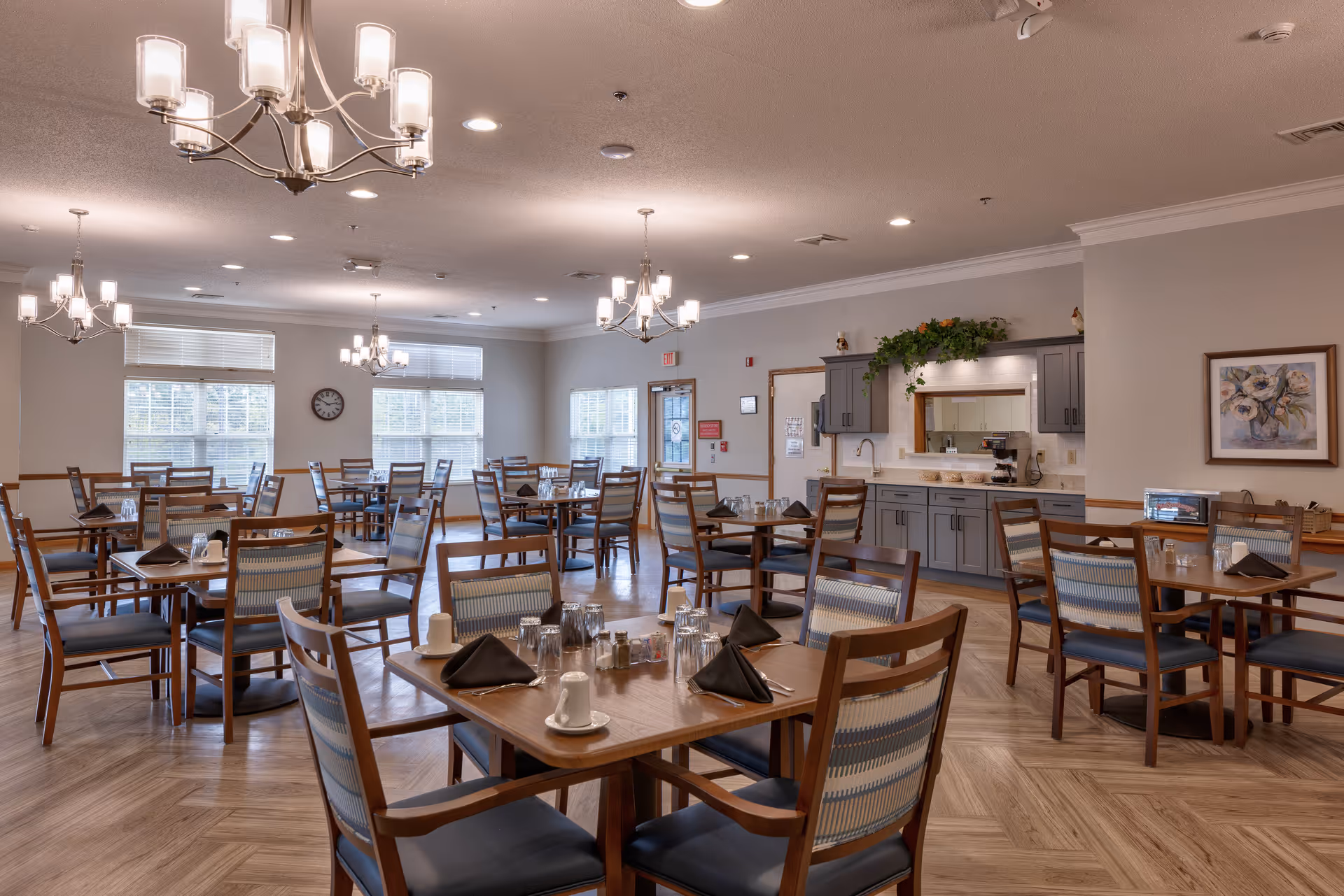 A spacious dining room with multiple wooden tables and chairs arranged neatly. Each table is set with black napkins, cups, and glasses. The room is well-lit with several modern chandeliers hanging from the ceiling. There are large windows allowing natural light to enter, and a counter area with cabinets and a coffee machine is visible along one wall. The walls are decorated with a framed painting and some greenery above the cabinets.