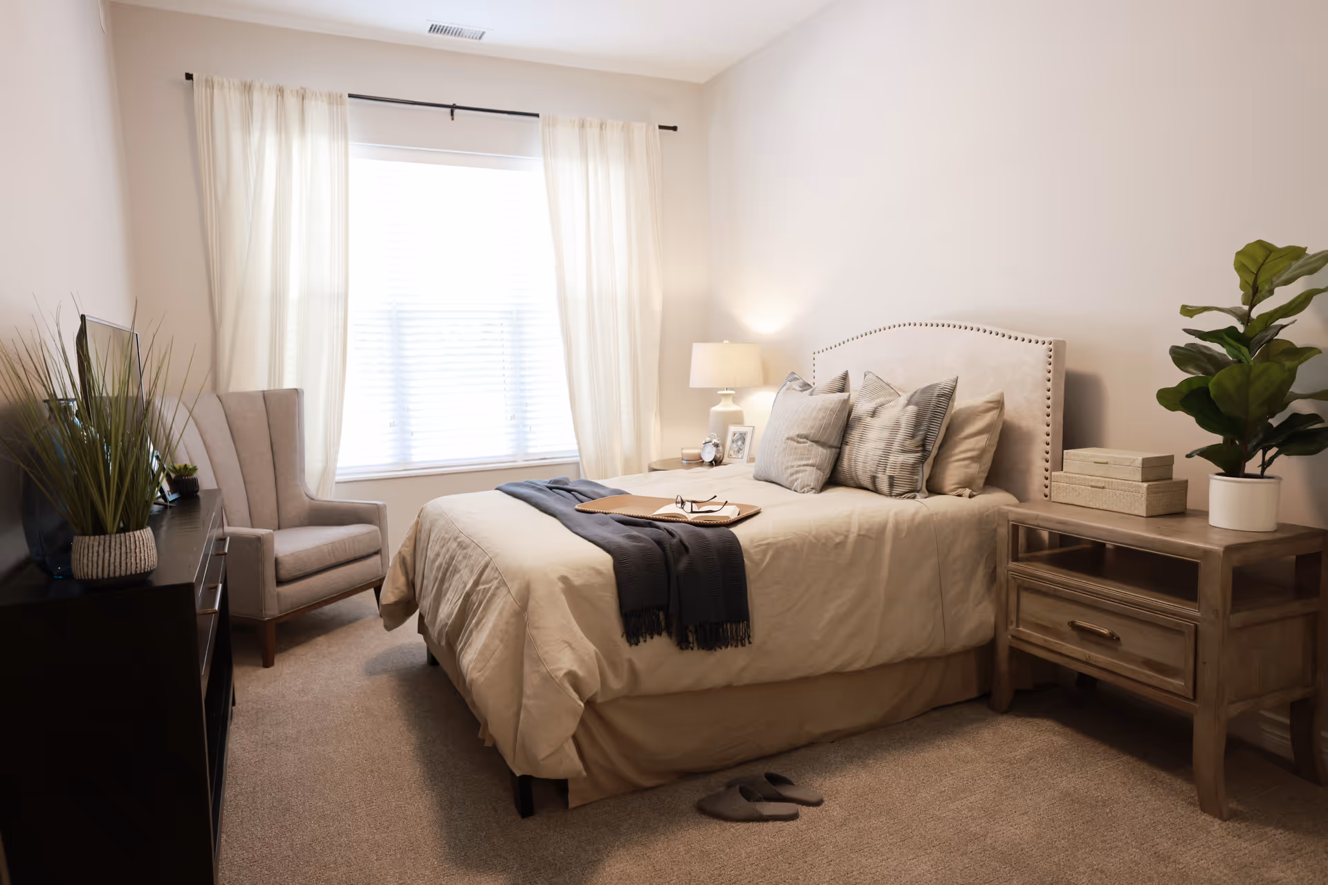 A cozy bedroom with a beige upholstered bed adorned with multiple pillows and a dark throw blanket. There is a wooden nightstand with a lamp, a plant, and decorative boxes on the right side of the bed. A comfortable armchair is placed near a window with sheer white curtains, and a dresser with a TV and plants is on the left side. The room has carpeted flooring and neutral-colored walls.