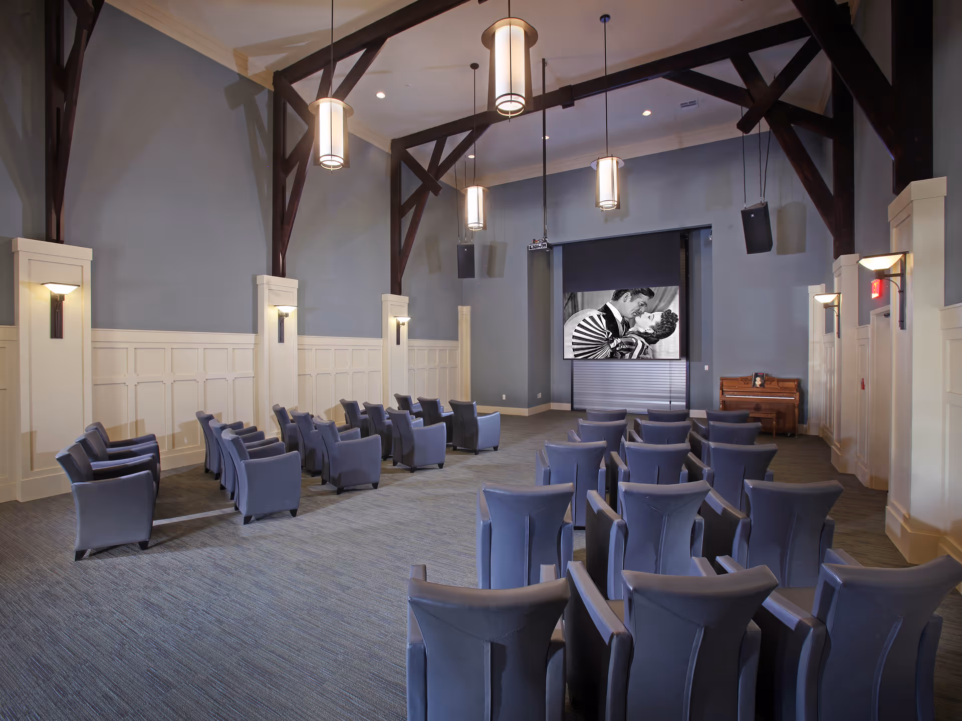 A large room with rows of blue upholstered chairs arranged facing a screen showing a black and white image of a couple kissing. The room has high ceilings with exposed dark wooden beams, modern hanging light fixtures, and wall sconces. There is a piano against the far wall to the right of the screen.