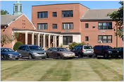 Exterior view of a multi-story brick building with several parked cars in front on a grassy lawn, under a clear sky.