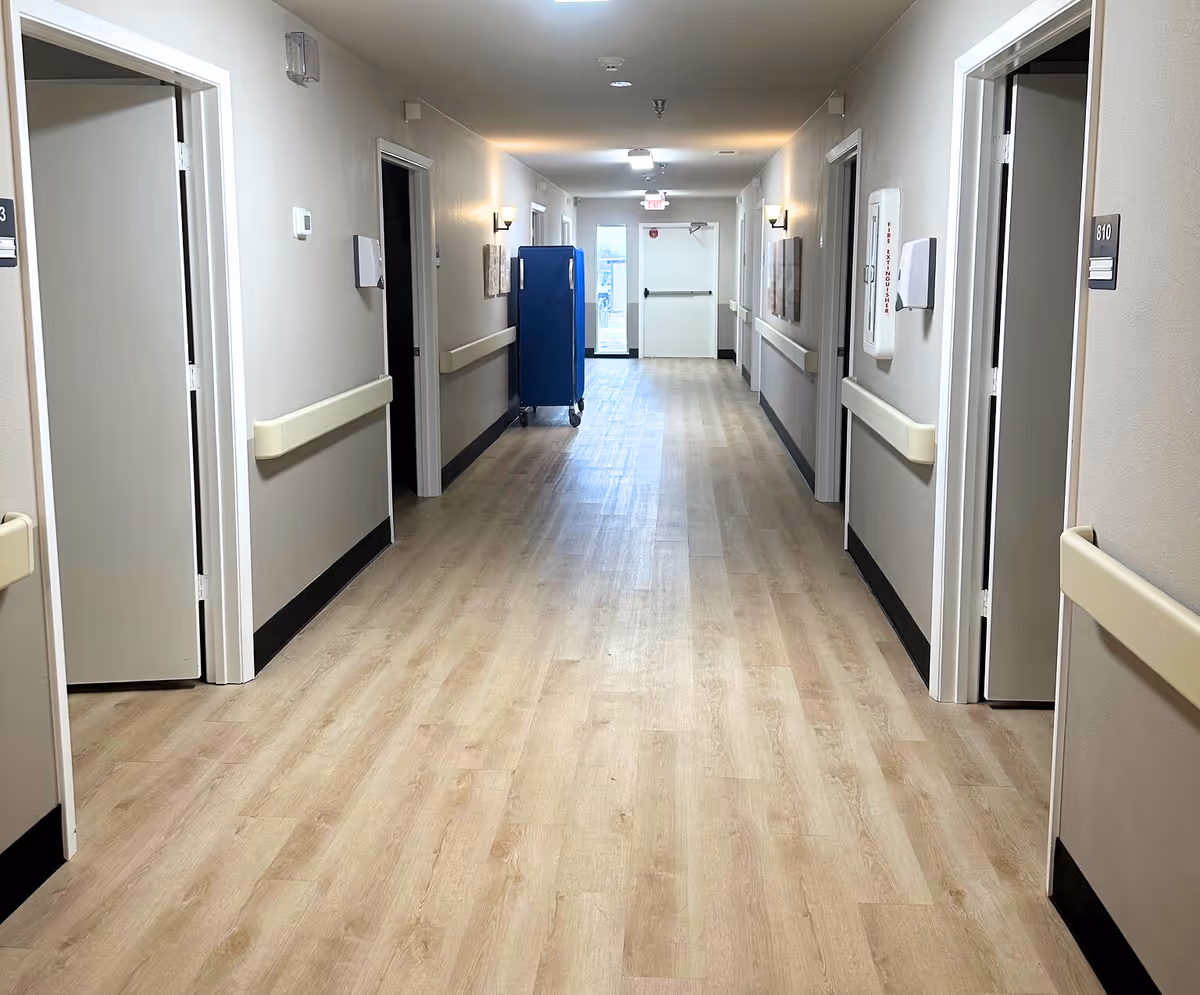 A clean, well-lit hallway in a nursing and rehabilitation facility with light wood flooring, beige walls, handrails on both sides, several open doors leading to rooms, wall-mounted lights, and a blue cart near the end of the corridor.