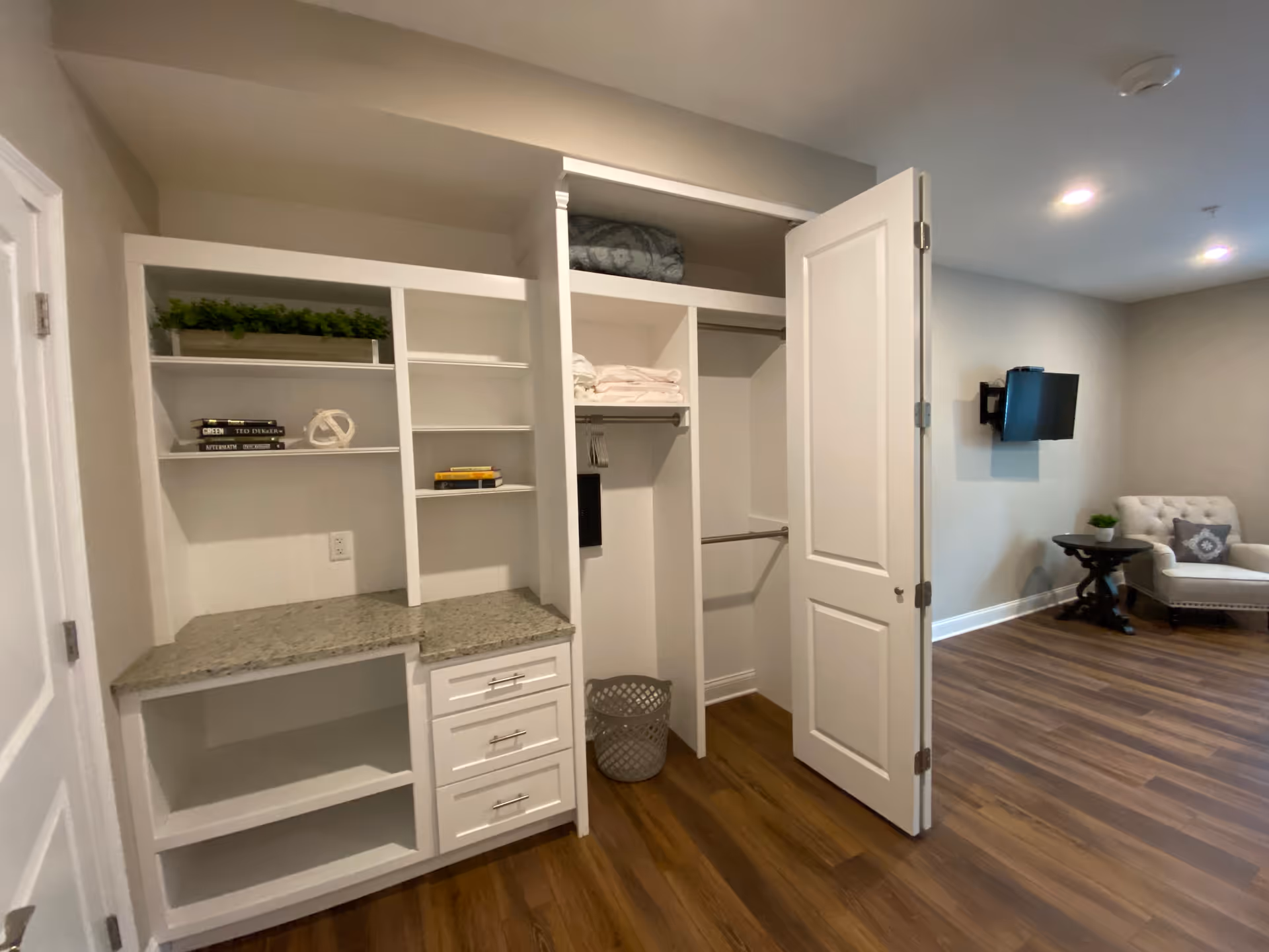 Interior view of a room featuring an open white closet with shelves, drawers, and hanging space. The closet contains folded towels, a laundry basket, and decorative items on the shelves. The room has wood flooring, a wall-mounted TV, a small round table with a plant, and a cushioned armchair with a decorative pillow.