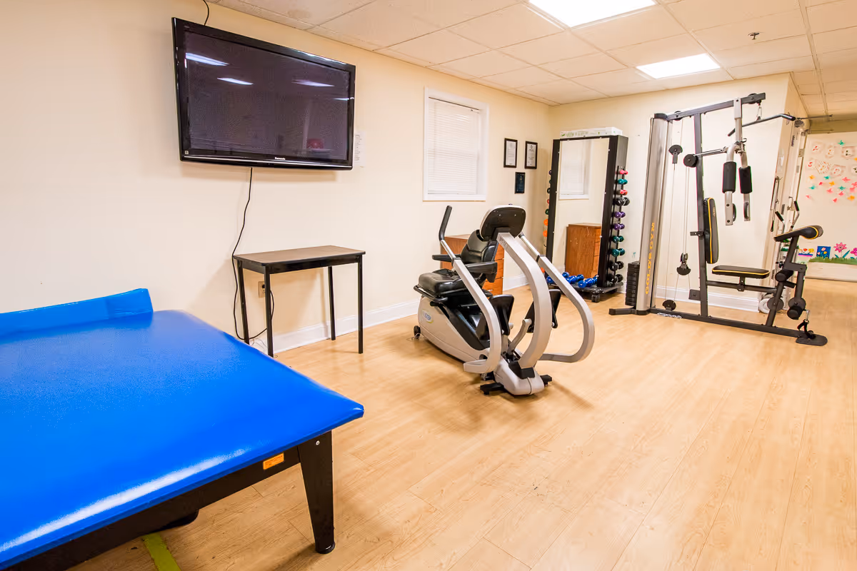 A rehabilitation center exercise room with a blue padded therapy table, a recumbent exercise bike, a multi-function weight machine, a rack of colorful dumbbells, a wall-mounted TV, and a small table against a beige wall with a window.